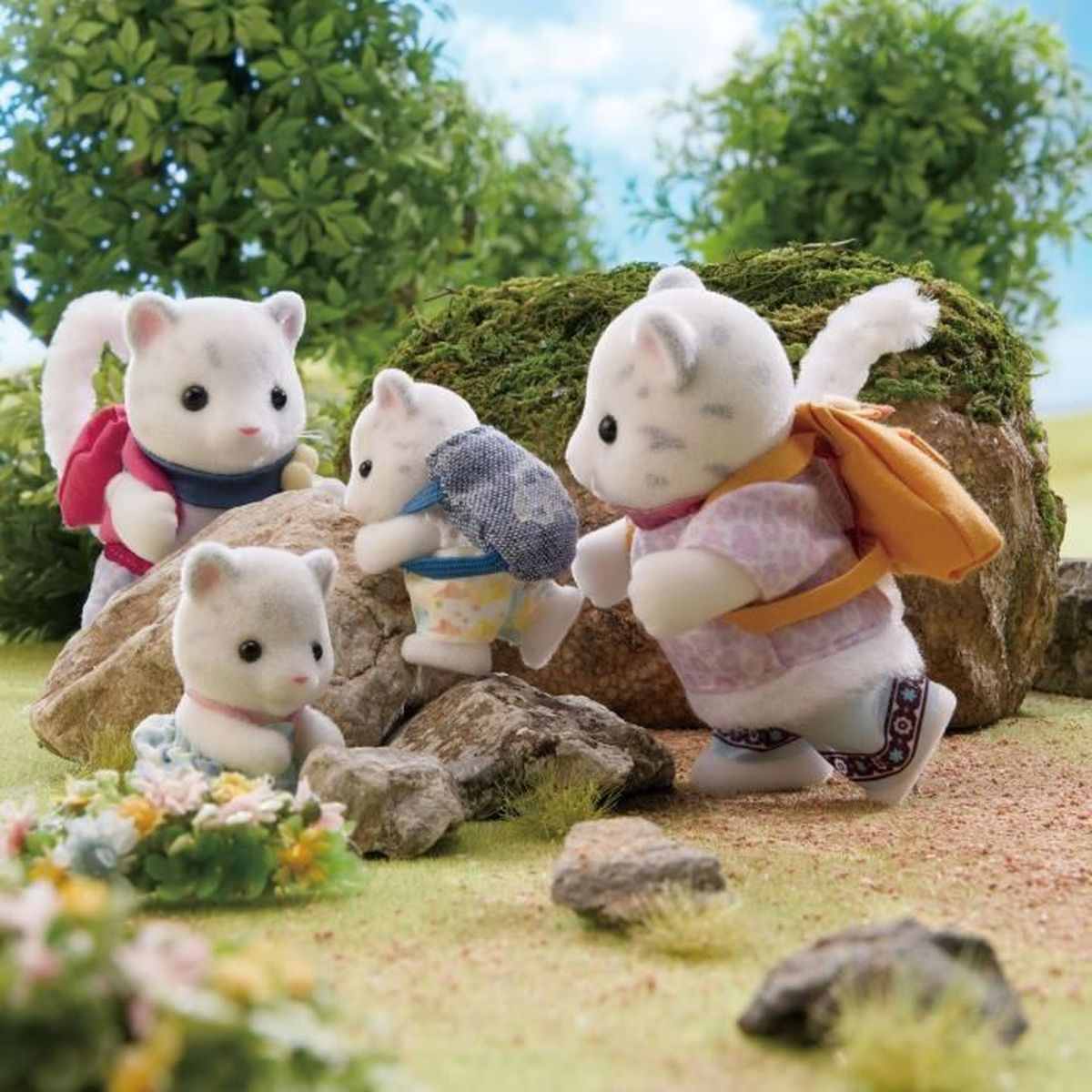 Cuatro figuras de gatos de juguete con mochilas están en un área cubierta de hierba con rocas. Follaje verde y cielo azul al fondo.