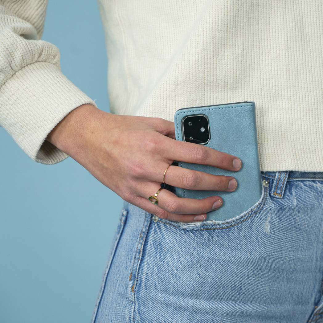 Una mano tiene una custodia blu per telefono in una tasca dei jeans. Un anello d'oro è sul dito. Lo sfondo è blu.