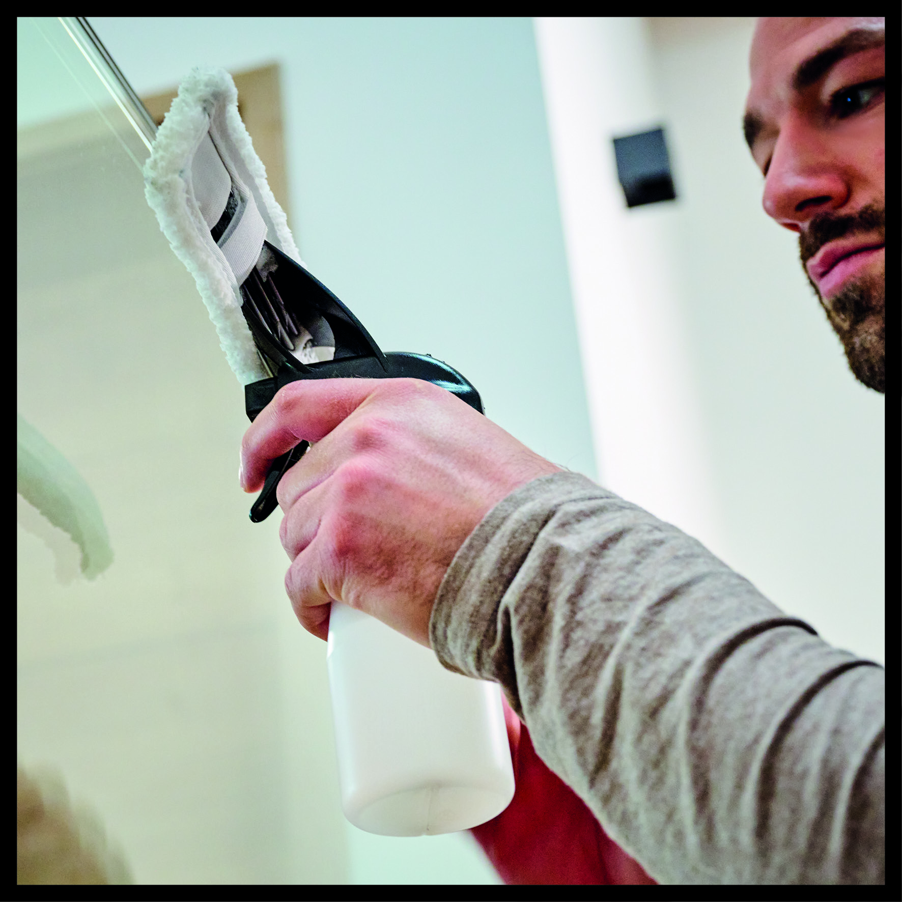 Mężczyzna czyści szklaną powierzchnię za pomocą ściągaczki i butelki z rozpylaczem. Ostrość skupiona jest na jego dłoni trzymającej narzędzia do czyszczenia.