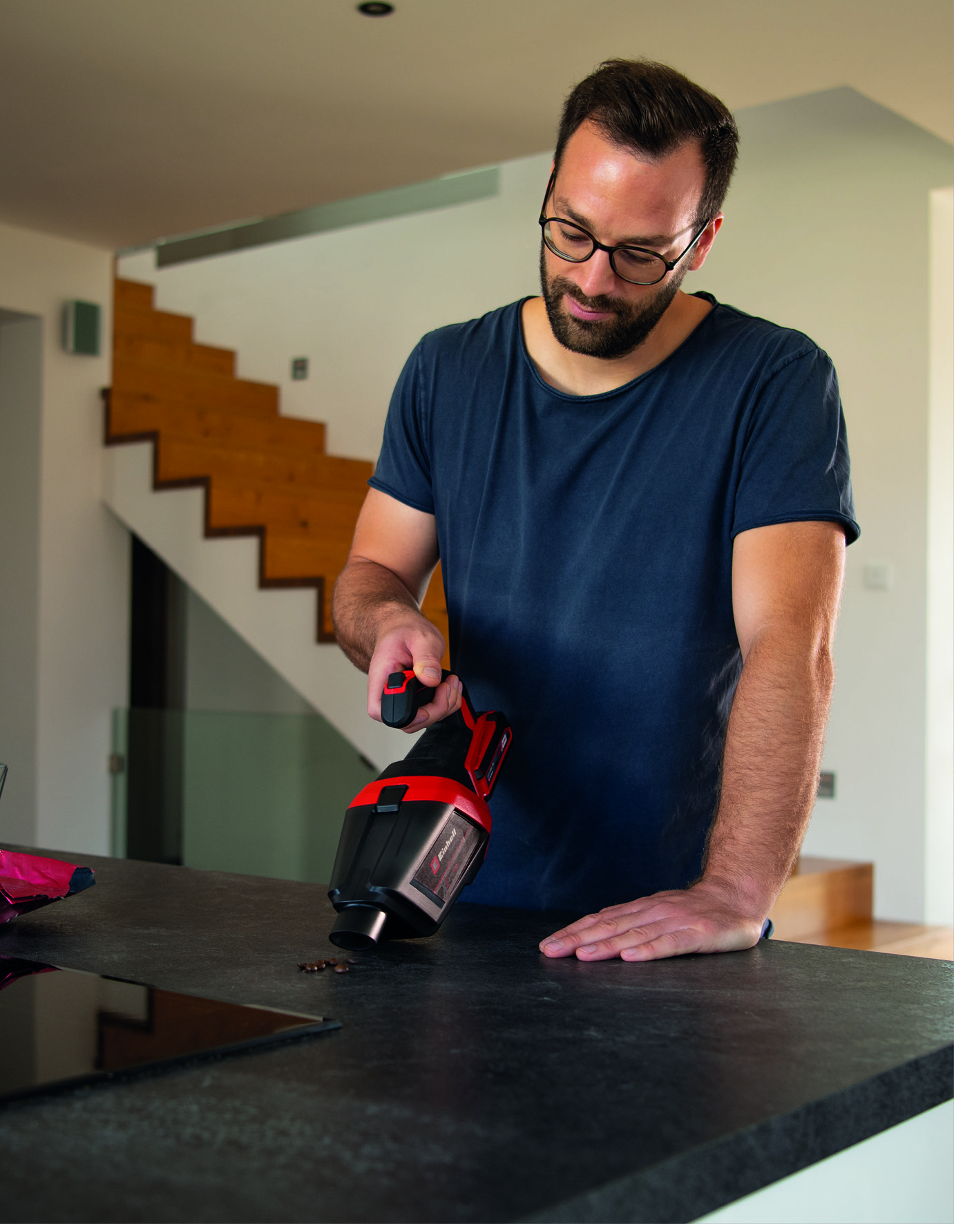 Ein Mann mit Brille reinigt mit einem Handstaubsauger eine Arbeitsplatte. Holztreppen sind im Hintergrund.