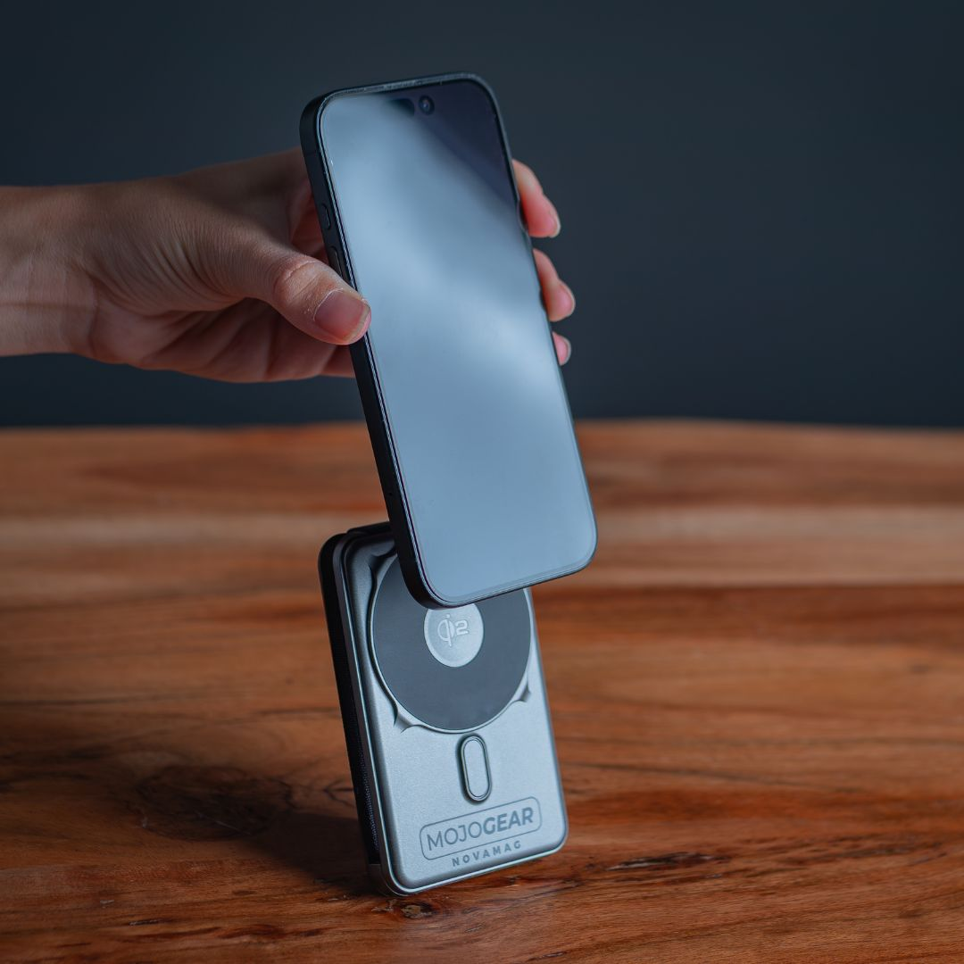 Eine Hand hält ein schwarzes Smartphone über einem silbernen MojoGear Novamag-Ladegerät auf Holz, dunkler Hintergrund.