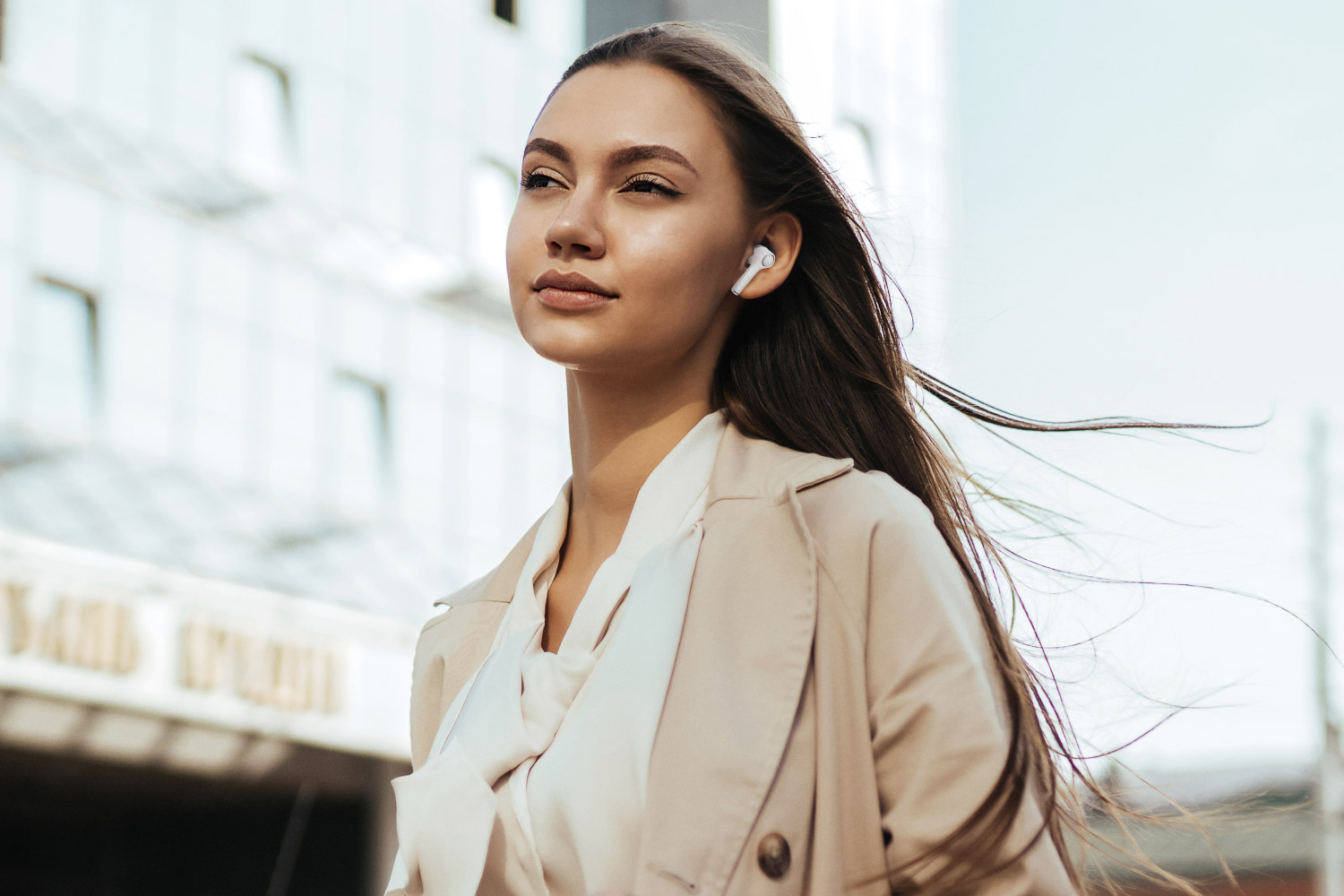 Kobieta z długimi brązowymi włosami w beżowym płaszczu i białej koszuli, z budynkiem w tle i słuchawkami.