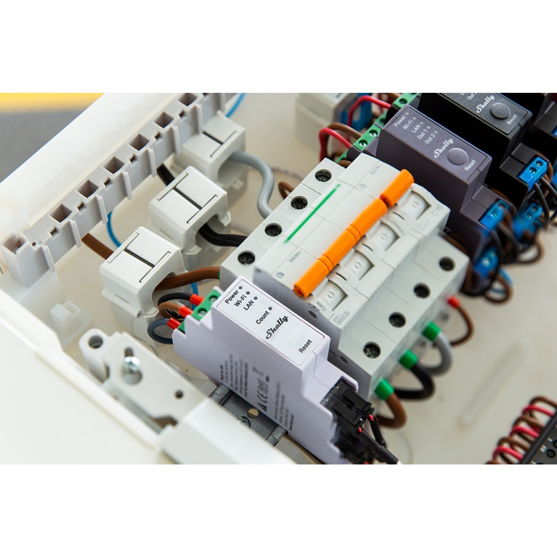 Panel elektryczny z różnymi komponentami, w tym wyłącznikami, przewodami i urządzeniem Shelly. Kolory to biały, pomarańczowy, czarny i zielony.