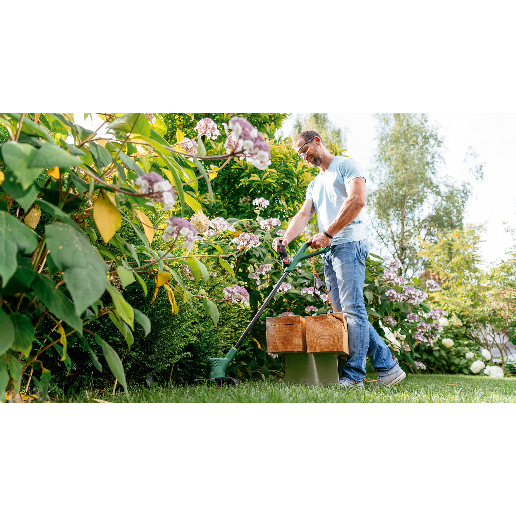 Ein Mann in blauem Hemd und Jeans benutzt einen Rasentrimmer in einem Garten. Blumen und Grün umgeben ihn.