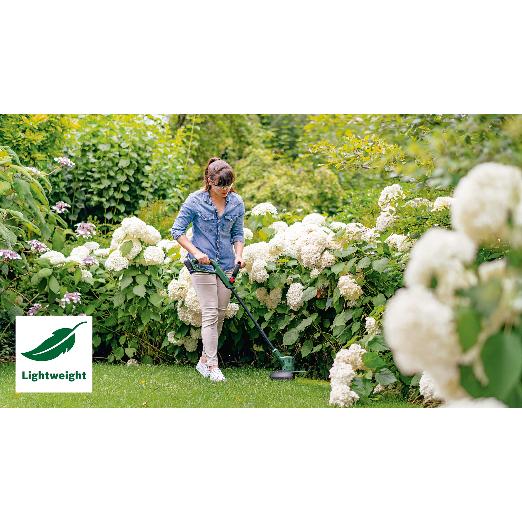 Eine Frau im Garten benutzt einen Rasentrimmer. Sie ist von weißen Blumen und grünen Büschen umgeben.