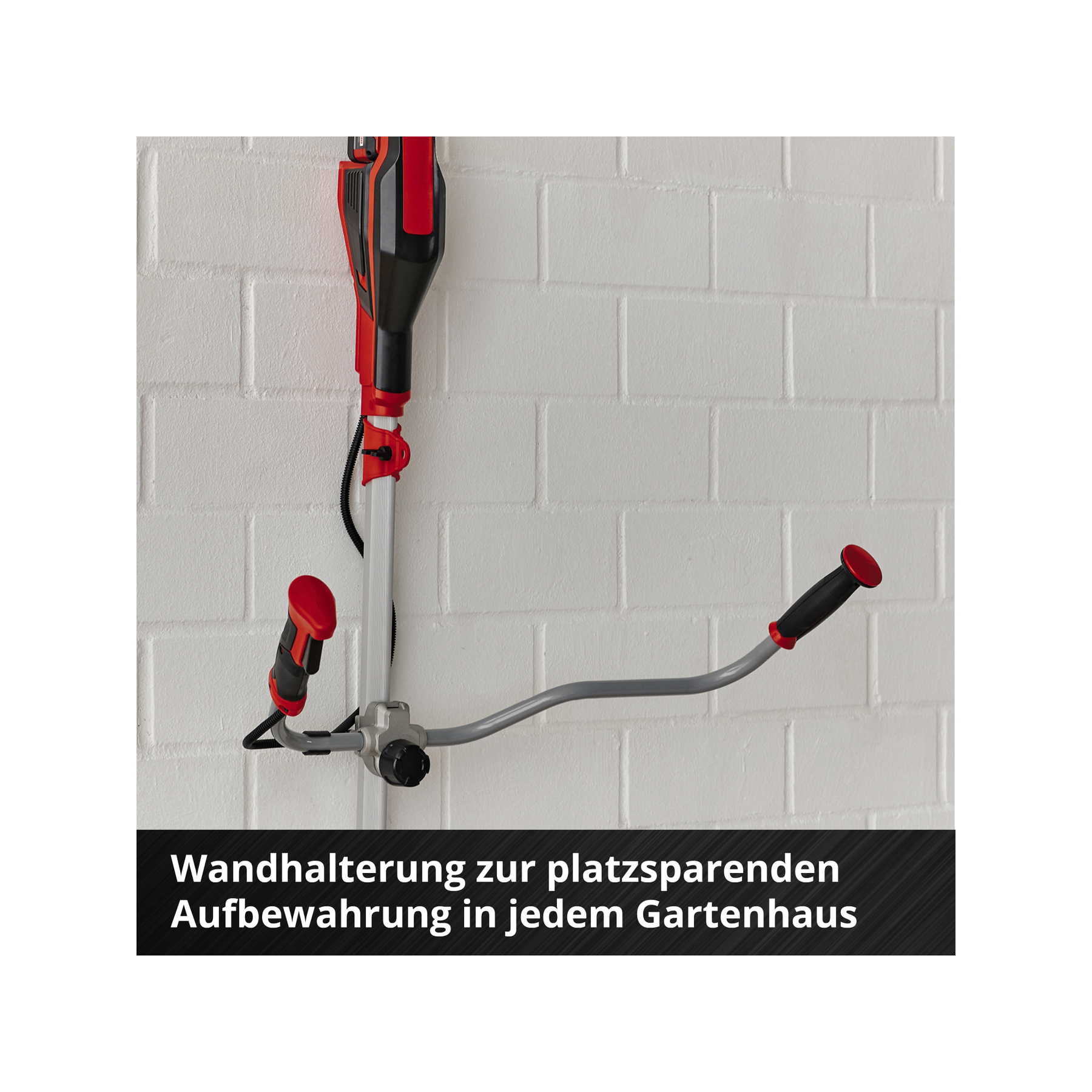Eine Wandhalterung hält ein rot/schwarzes Werkzeug. Die Halterung ist grau, mit roten Griffen, an einer weißen Ziegelmauer montiert.