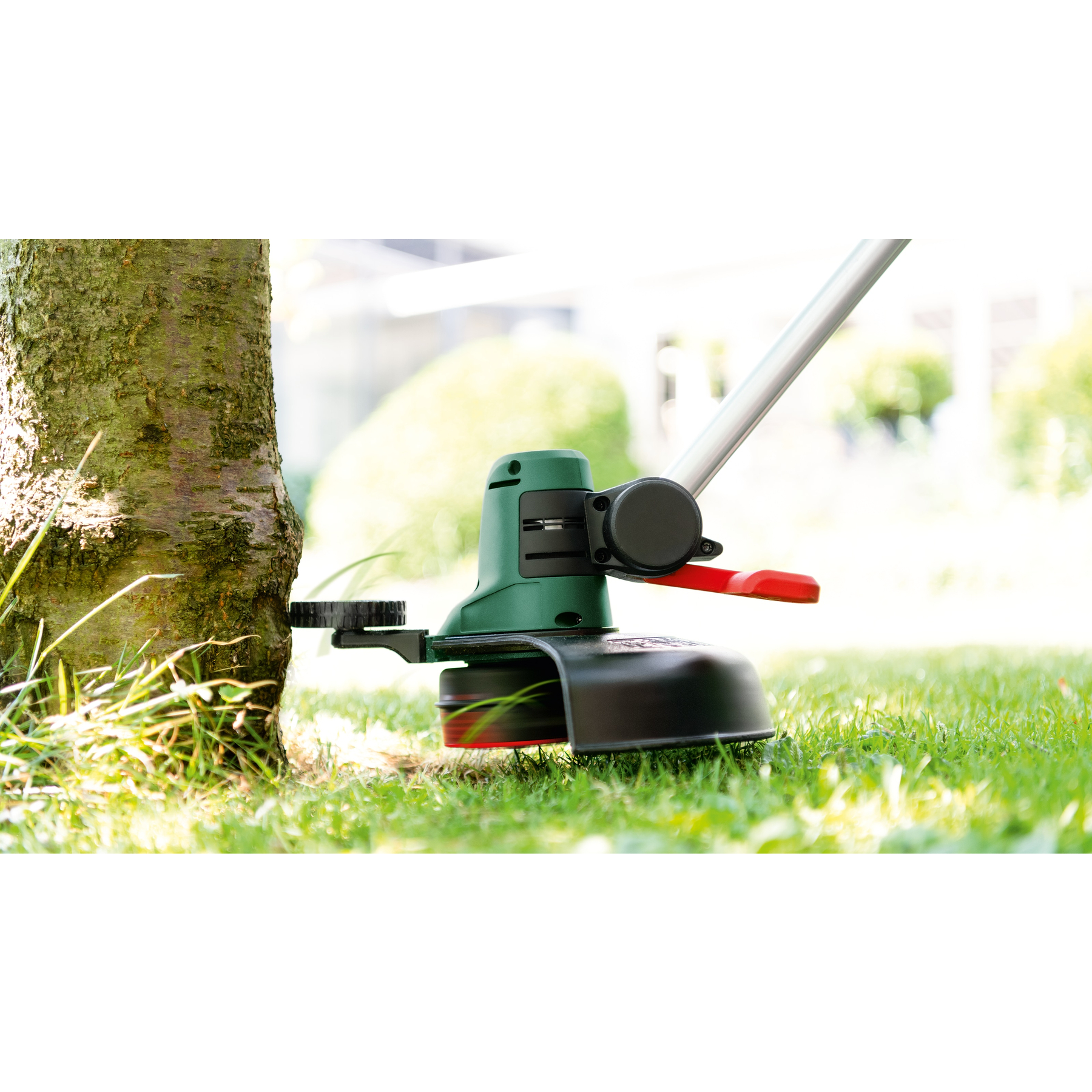 Ein grüner und schwarzer Rasentrimmer steht neben einem Baum. Der Hintergrund umfasst Gras und ein verschwommenes Gebäude.