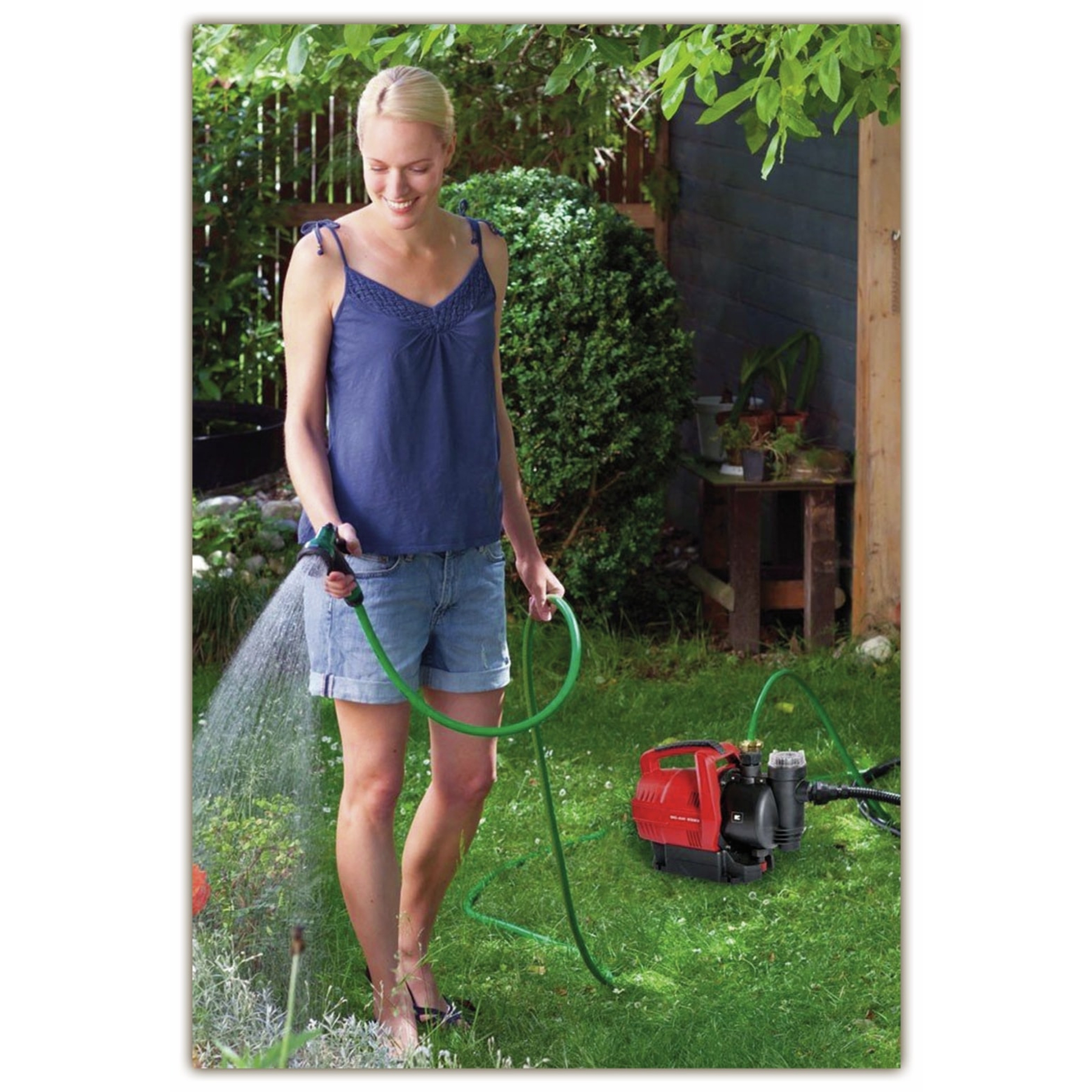 Kobieta podlewa rośliny w ogrodzie wężem. Czerwona pompa stoi na trawie. Widoczne zielone liście i drewniana konstrukcja.