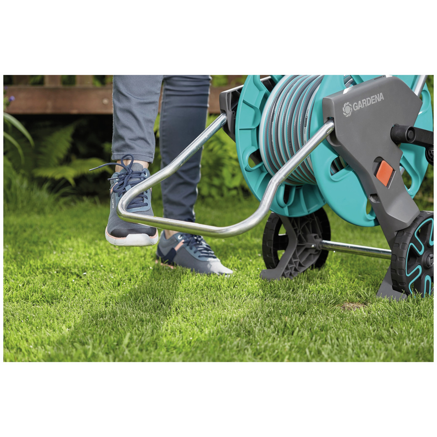 Person mit blauen Schuhen steht auf grünem Gras neben einer Gardena Schlauchtrommel. Schlauch ist türkis und grau. Hintergrund ist ein Holzdeck.