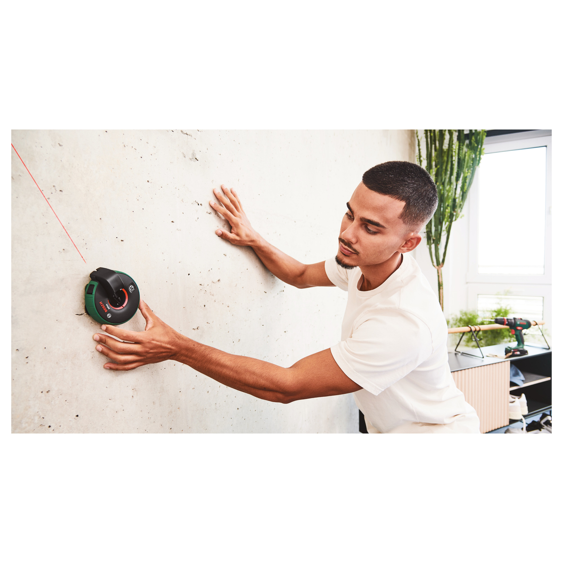 Ein Mann benutzt einen Laser auf einer Wand. Der Laser projiziert eine rote Linie. Eine Bohrmaschine und Pflanzen sind im Hintergrund.