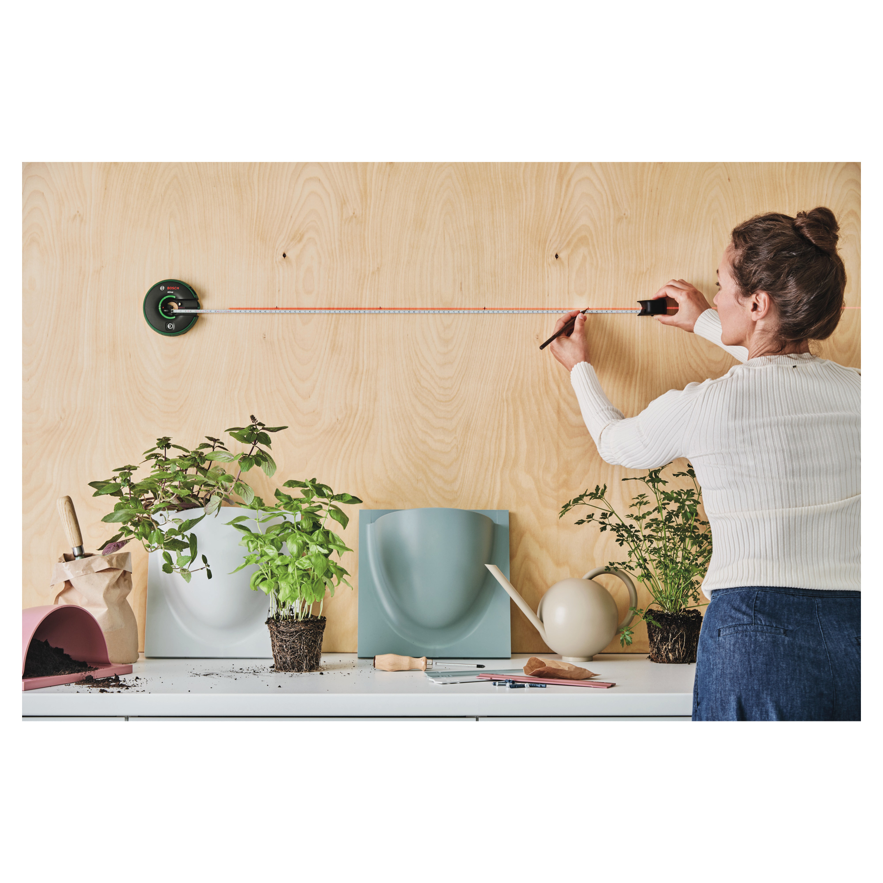 Frau misst an einer Holzwand mit Pflanzen und Gartengeräten davor. Ein Maßband wird verwendet.
