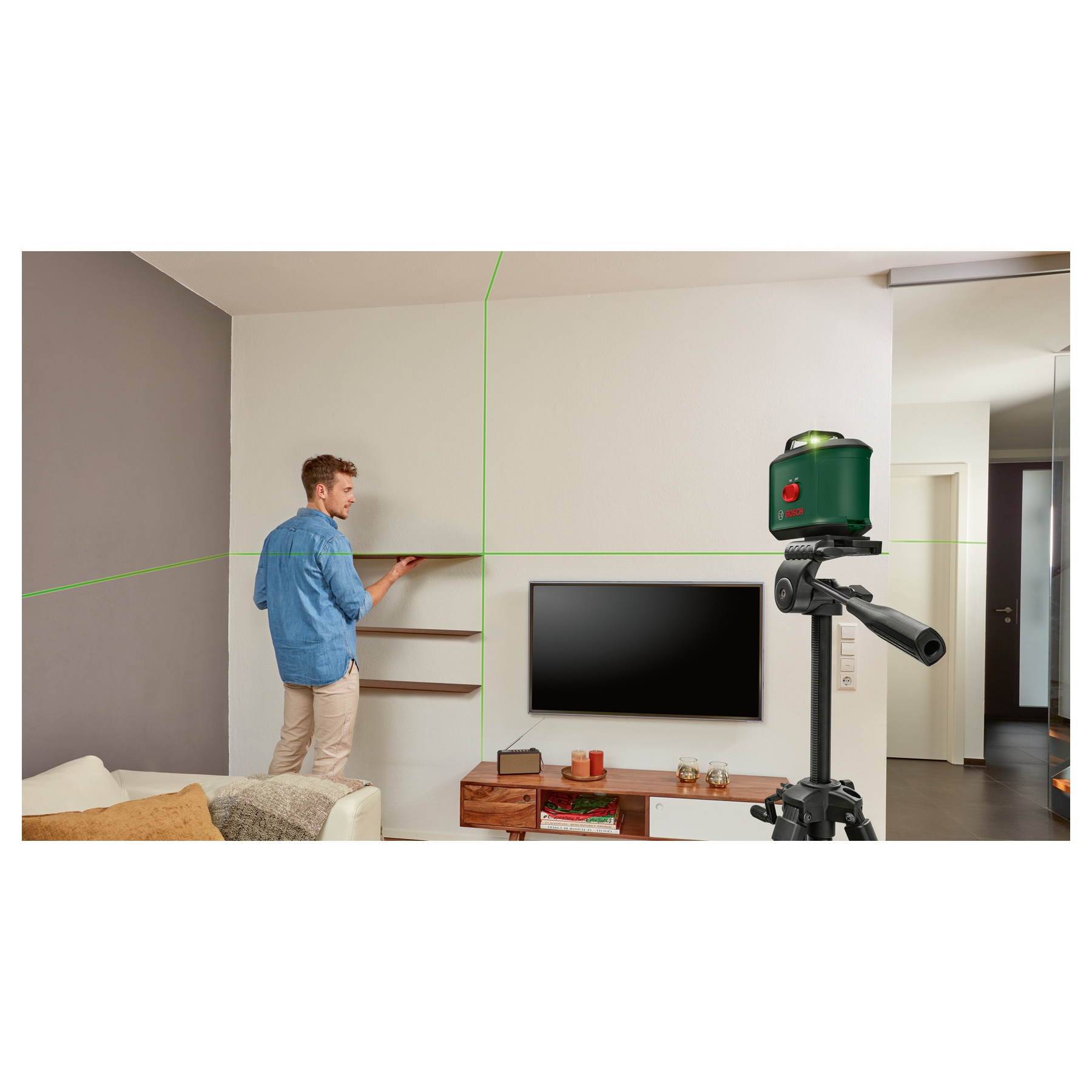 Mężczyzna instaluje półki za pomocą poziomicy. Zielony laser wyświetla linie. Widoczny jest telewizor, sofa i drewniane meble.