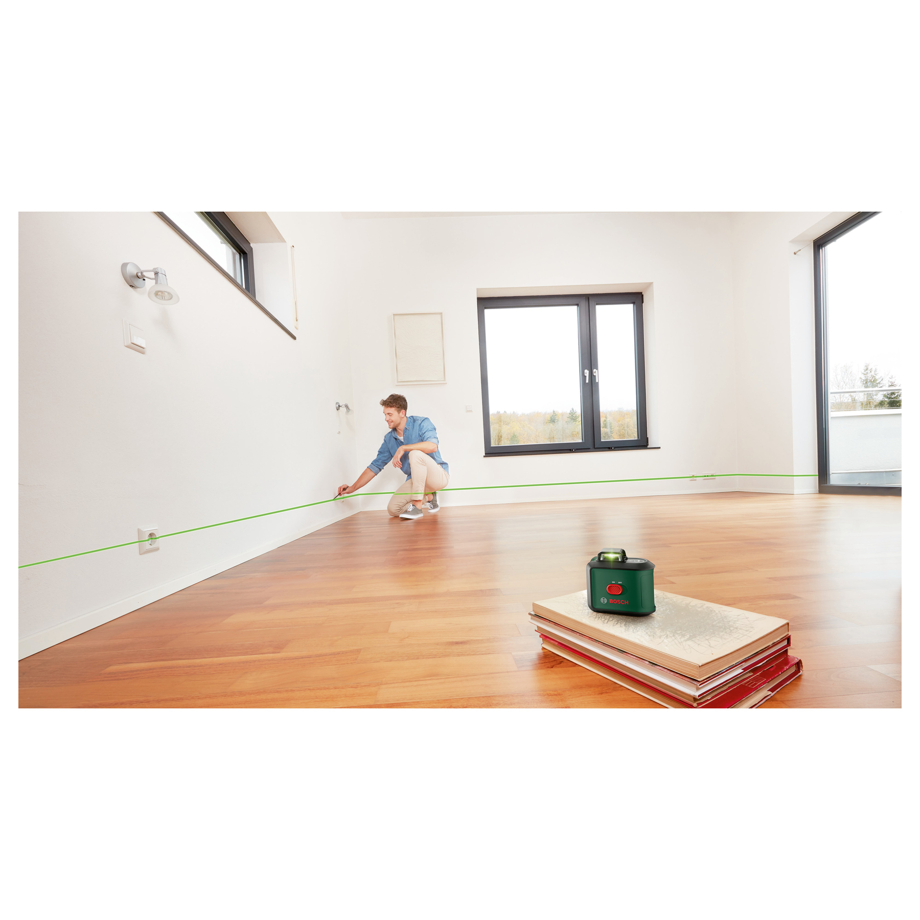 Mężczyzna używa poziomicy laserowej w pokoju. Zielone linie na ścianach i podłodze. Poziomica na książkach. Duże okno.