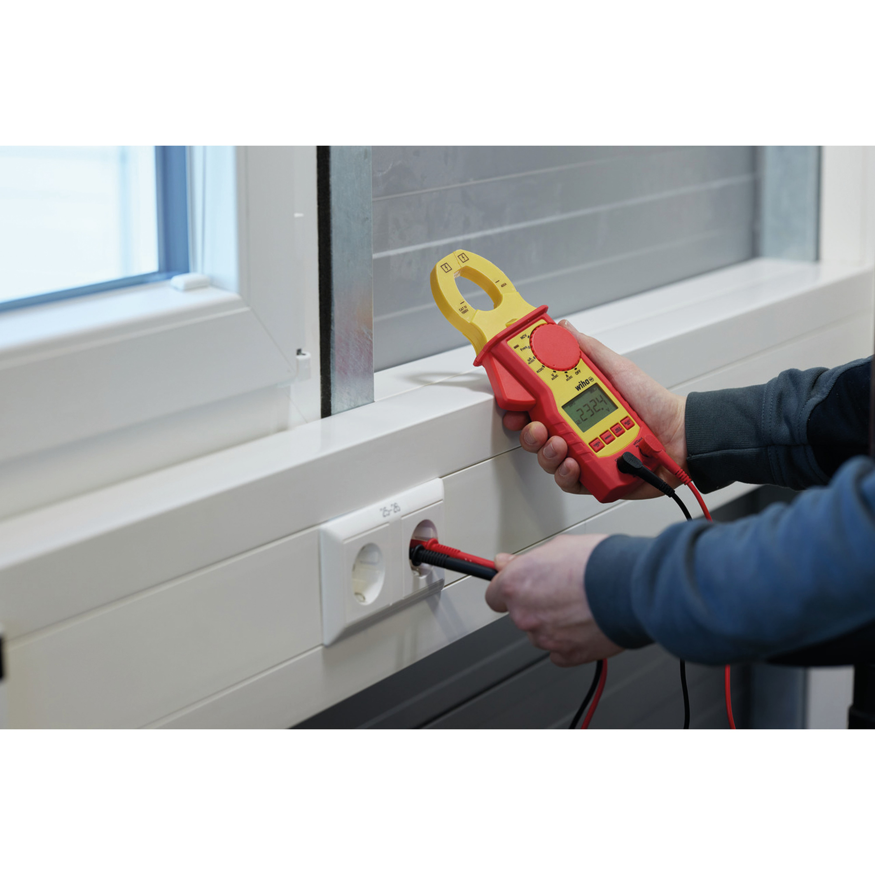 Eine Person verwendet ein rot-gelbes Zangenmessgerät, um die Spannung in einer Steckdose zu messen. Im Hintergrund ist ein Fenster.