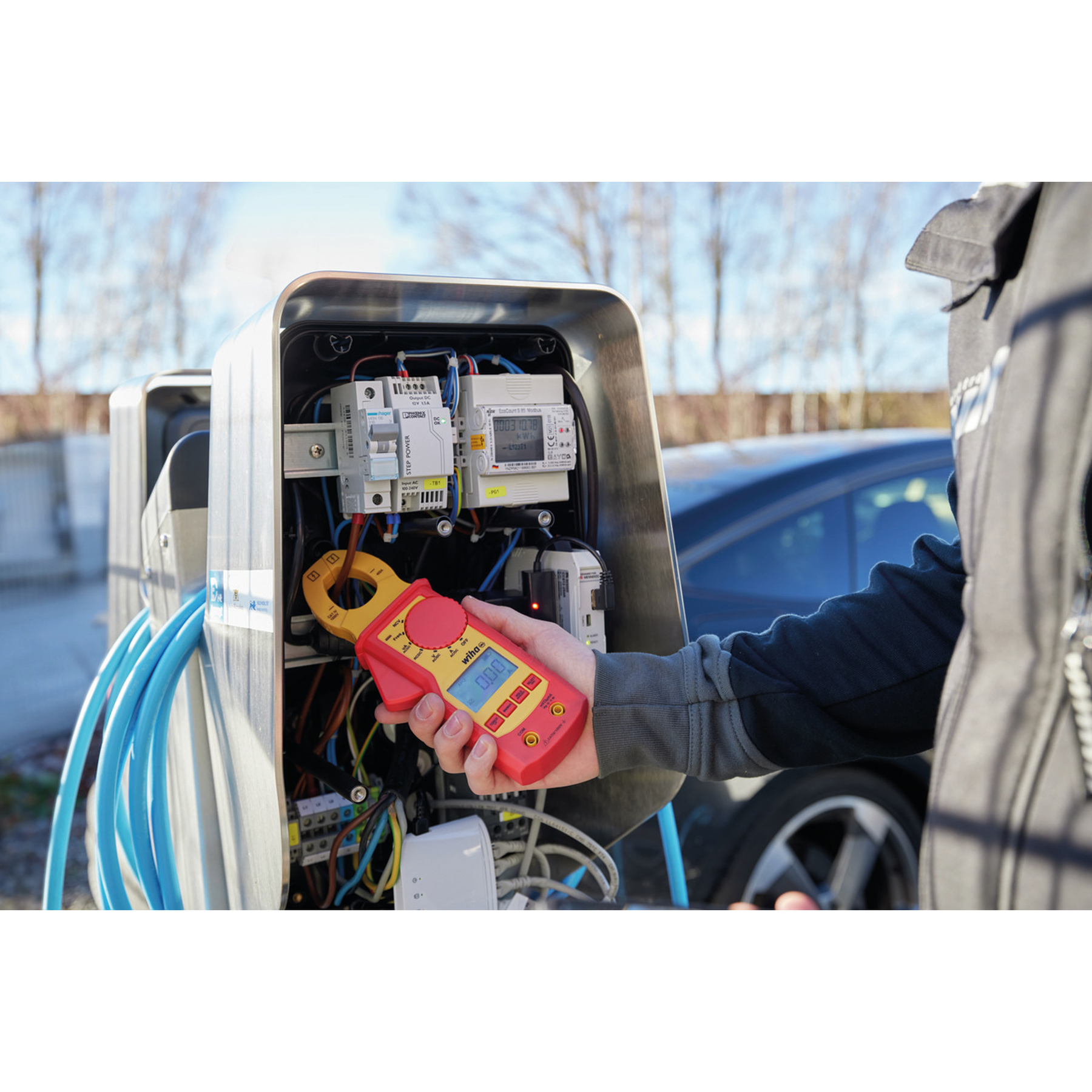 Eine Person verwendet ein Stromzangenmessgerät, um elektrische Komponenten in einer Ladestation zu überprüfen, mit einem Auto im Hintergrund.