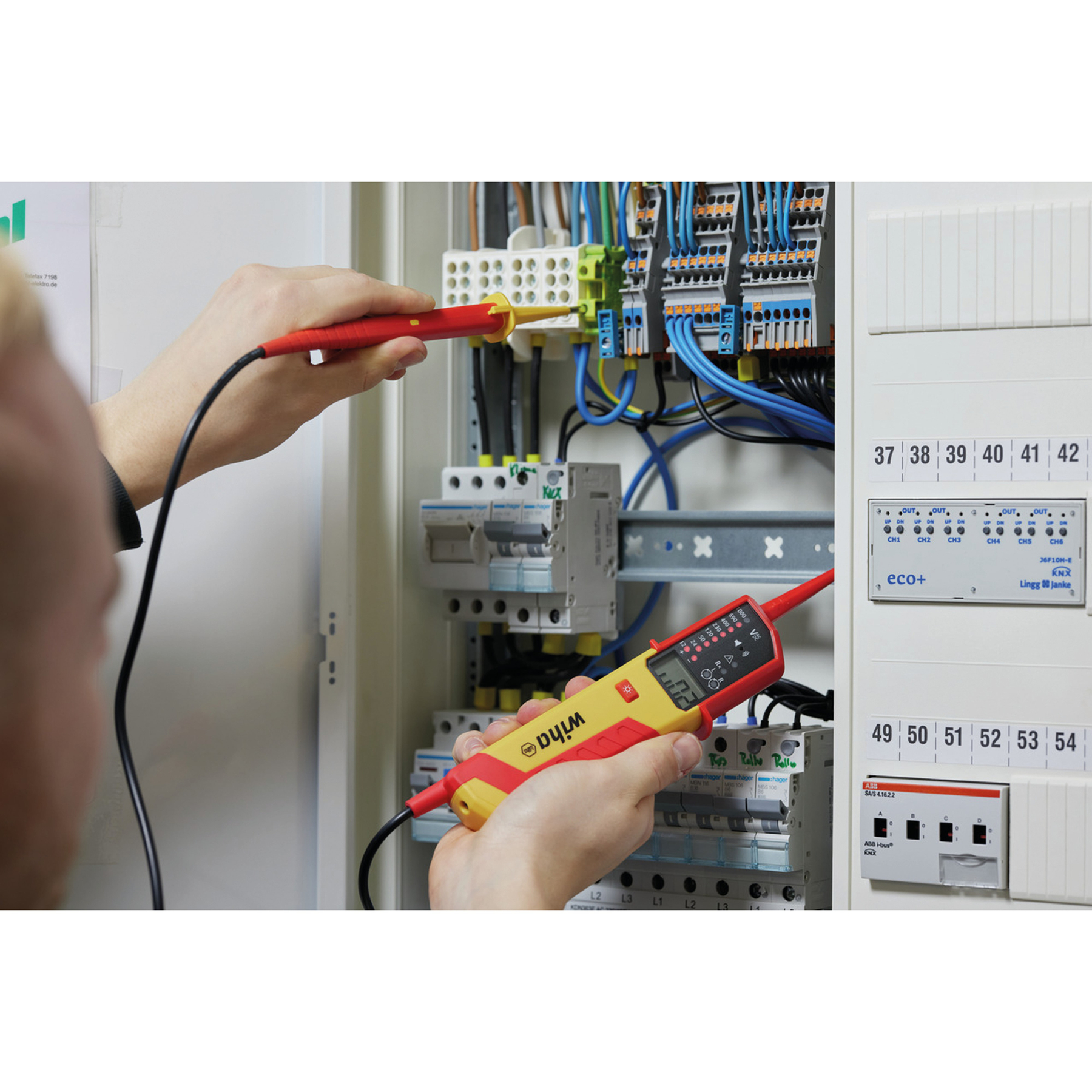 Eine Person verwendet einen roten und gelben Wiha-Spannungsprüfer an elektrischen Leitungen in einem Panel.