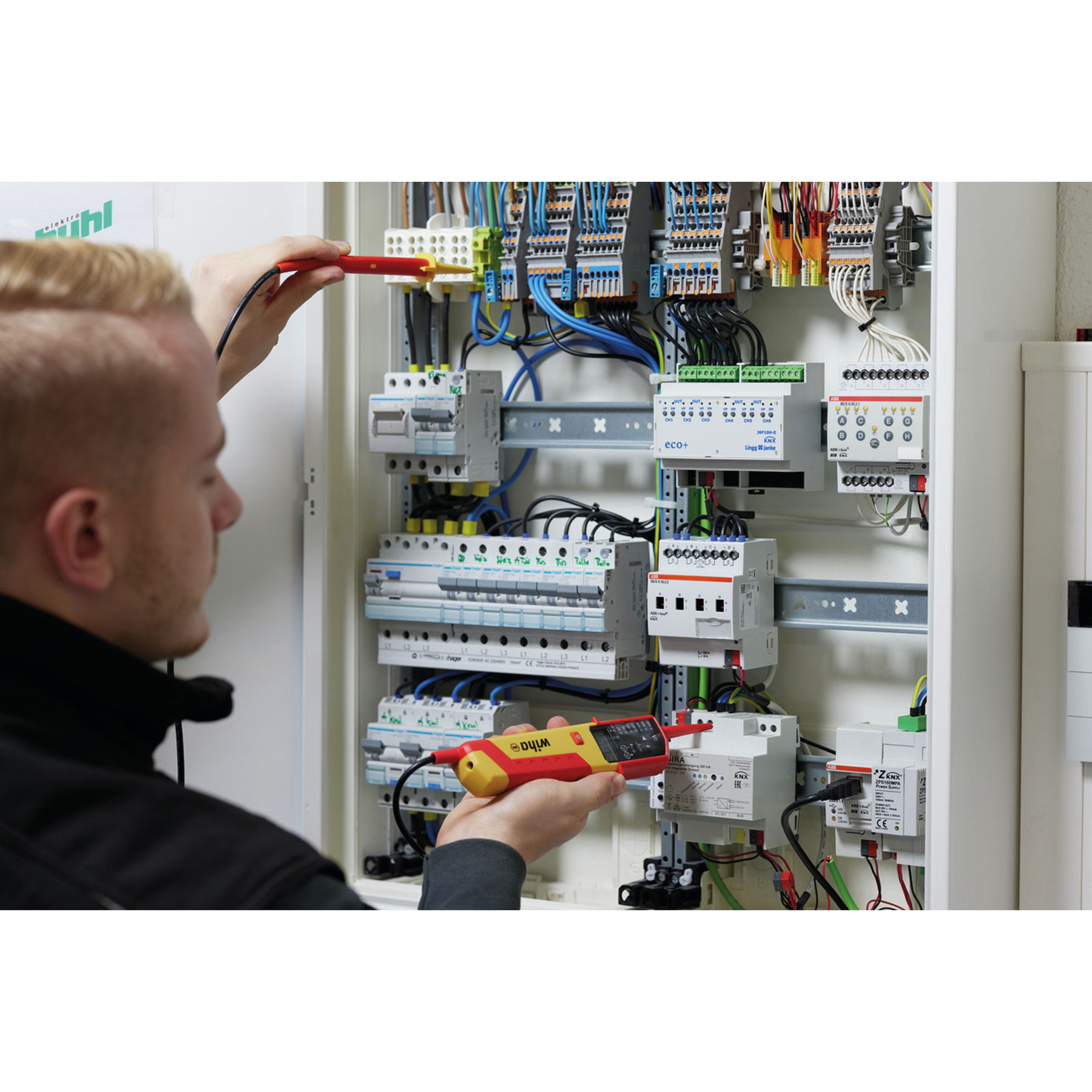 Ein Elektriker testet einen Sicherungskasten mit einem rot-gelben Spannungstester. Drähte und elektronische Bauteile füllen die Box.