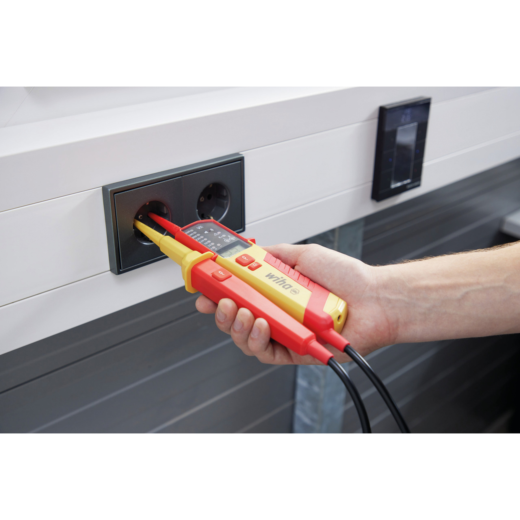Hand hält zwei elektrische Wiha-Tester in einer Steckdose. Die Tester sind rot und gelb. Hintergrund: weiße Wand und schwarzer Schalter.