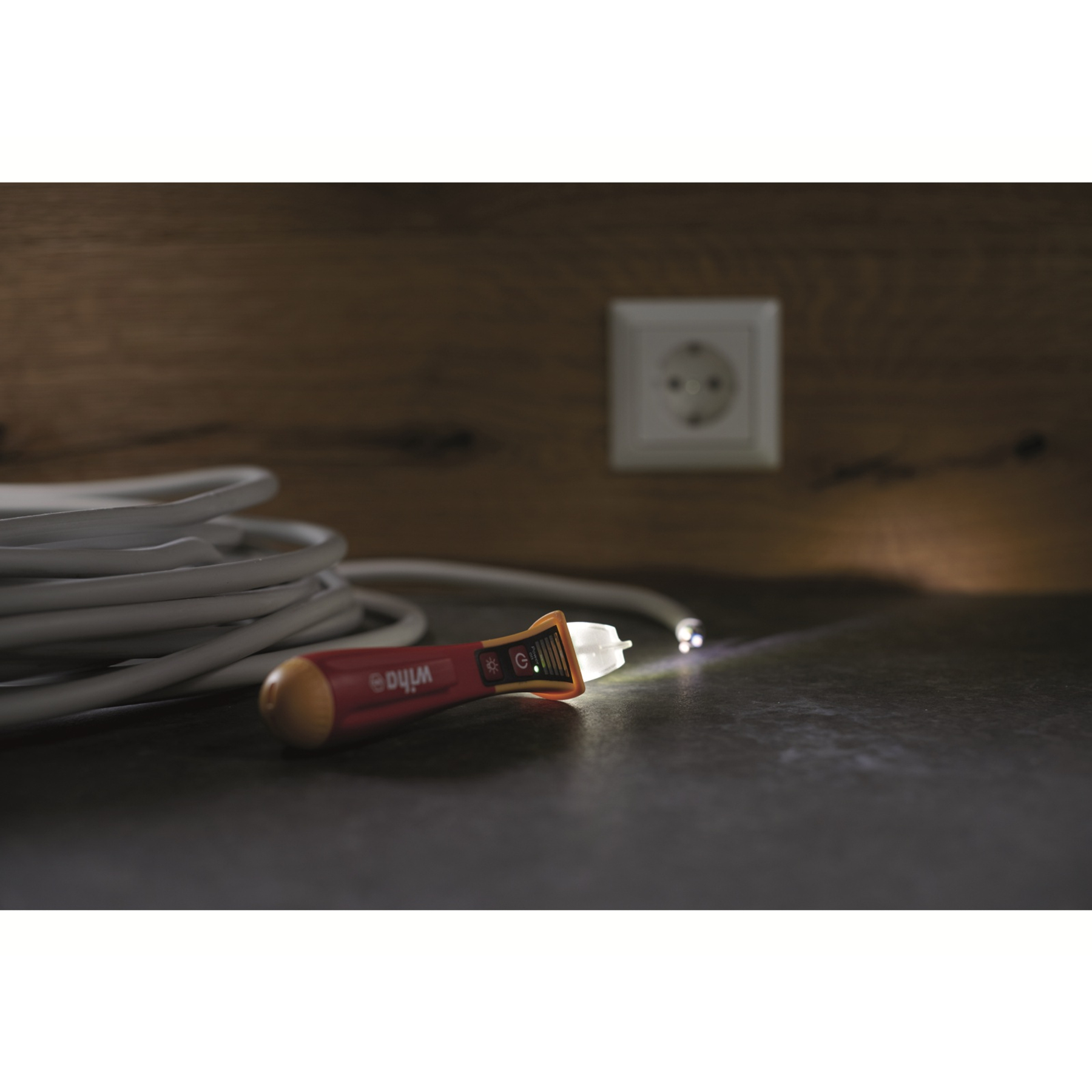 Czerwony tester napięcia leży na ciemnej powierzchni. Biały kabel i białe gniazdo w tle. Tester jest włączony.