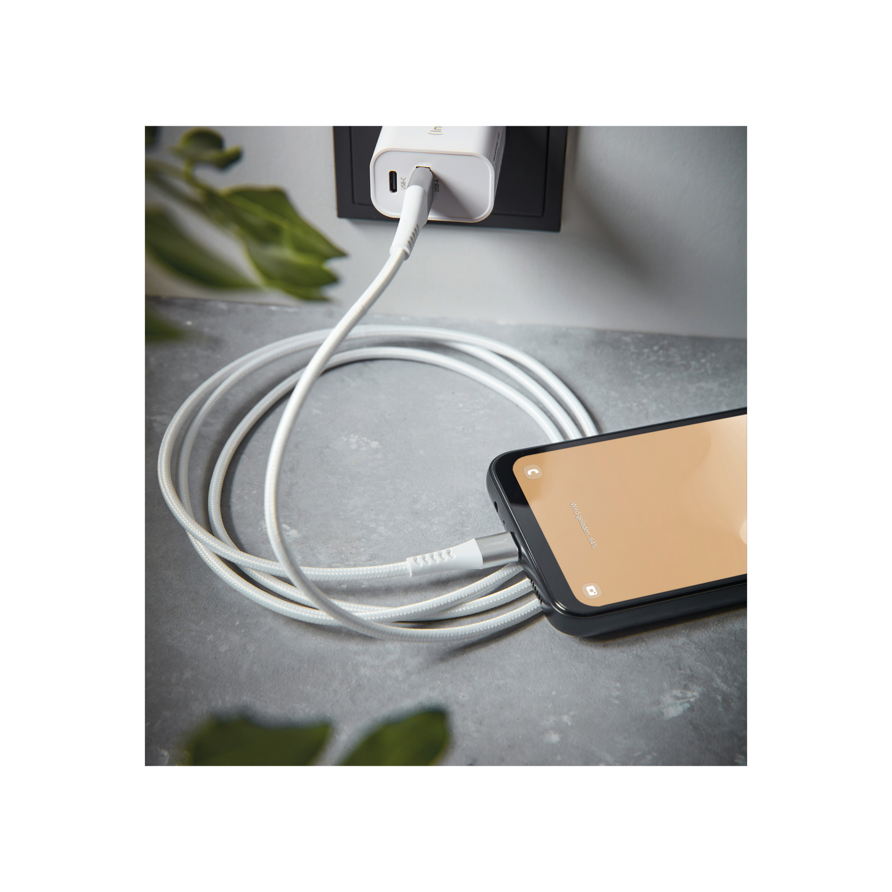 Un téléphone se charge avec un câble blanc. Le chargeur est branché au mur. L'écran du téléphone est visible.