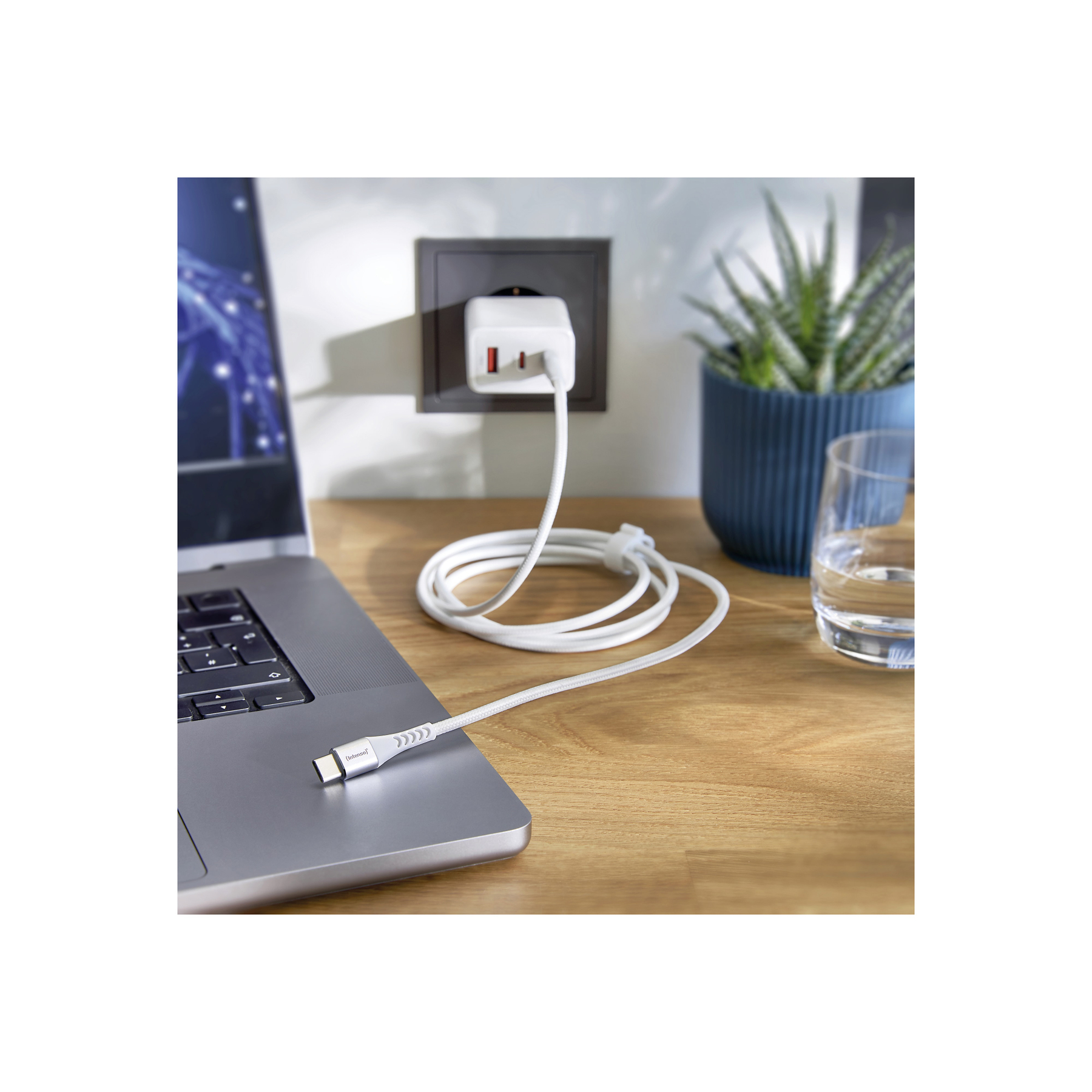 Un laptop collegato a un caricabatterie bianco tramite un cavo bianco. Un caricabatterie bianco è collegato a una presa a muro. Una pianta e un bicchiere sono visibili.