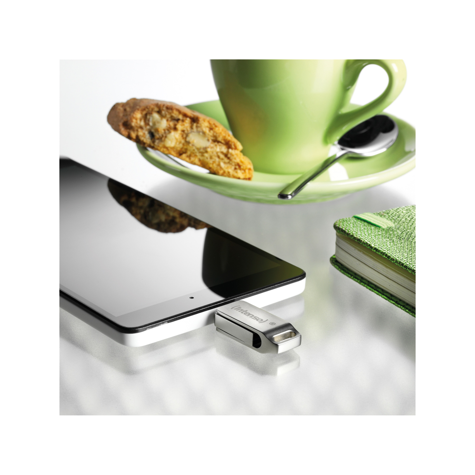 Tablet, pendrive, zielony kubek ze spodkiem, biscotti i zielony notatnik leżą na białej powierzchni.