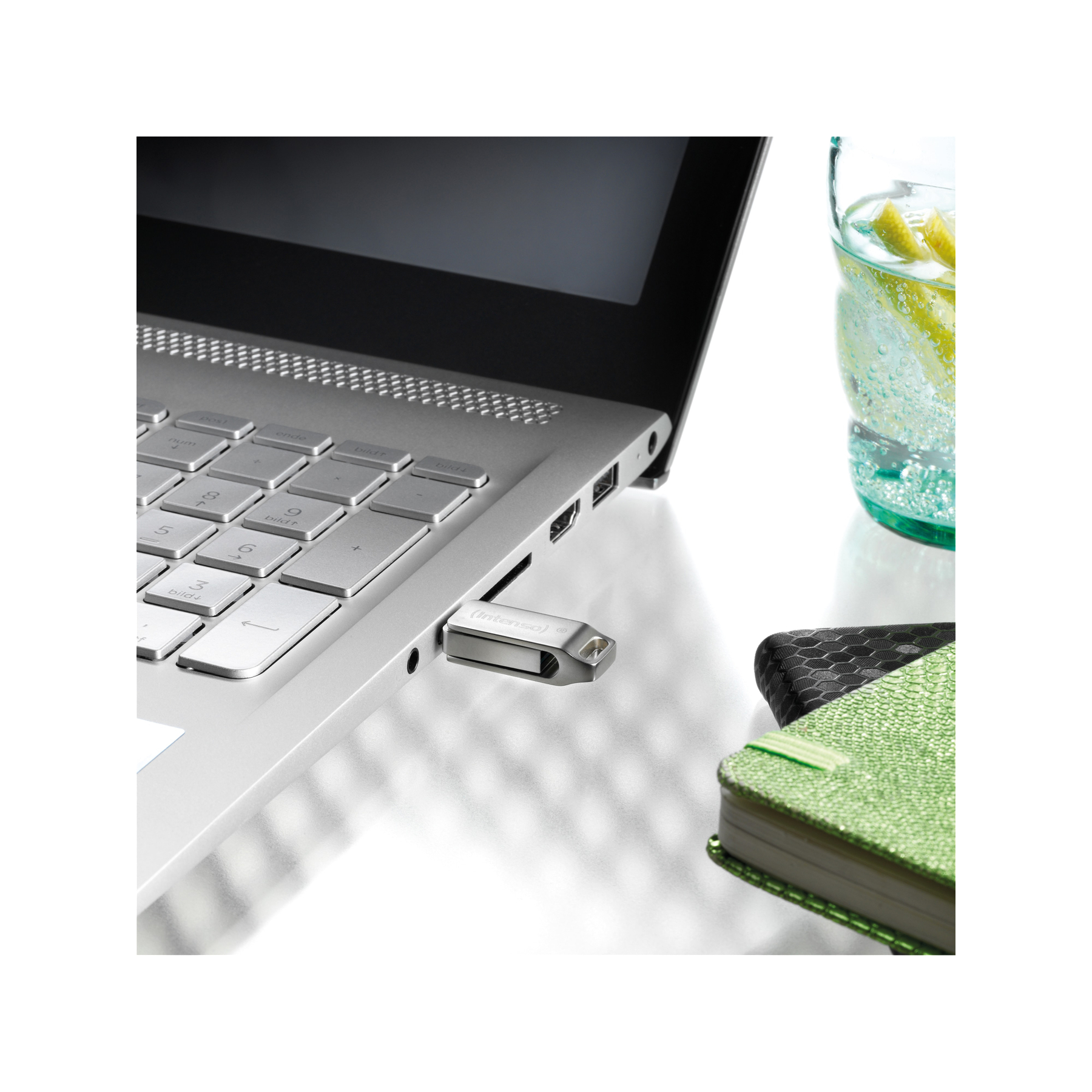 Laptop z włożonym pendrivem. Szklanka wody z plasterkami cytryny. Zielony notatnik obok.