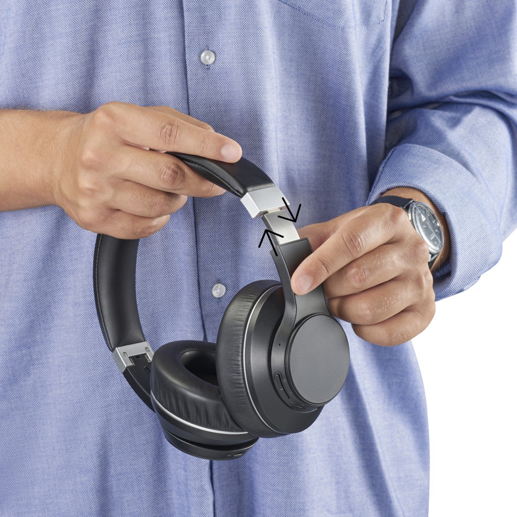 Osoba trzyma czarne słuchawki. Słuchawki są regulowane. Osoba ma na sobie niebieską koszulę i zegarek.