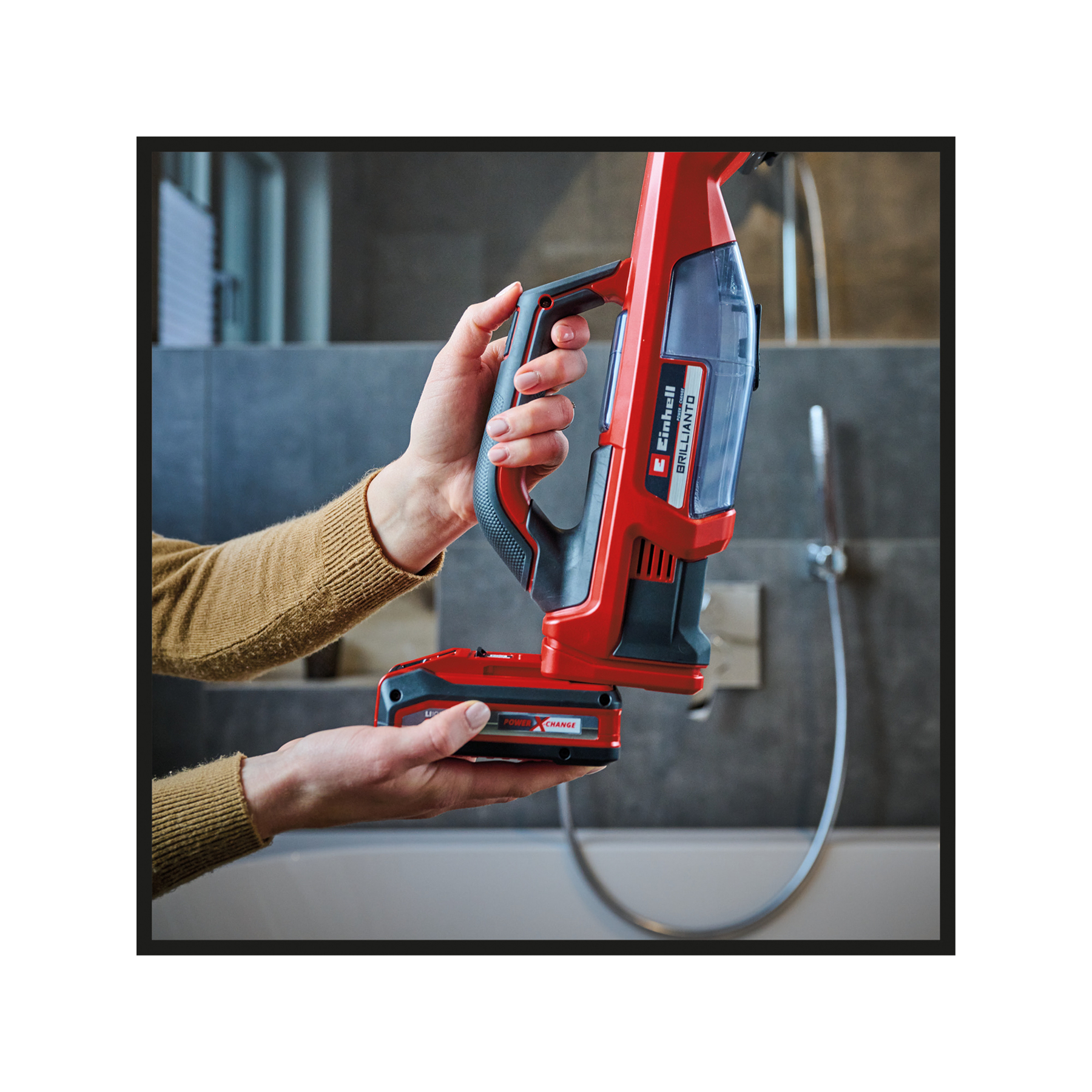 Ręce trzymają czerwono-czarny bezprzewodowy odkurzacz. Odkurzacz jest na tle łazienki.