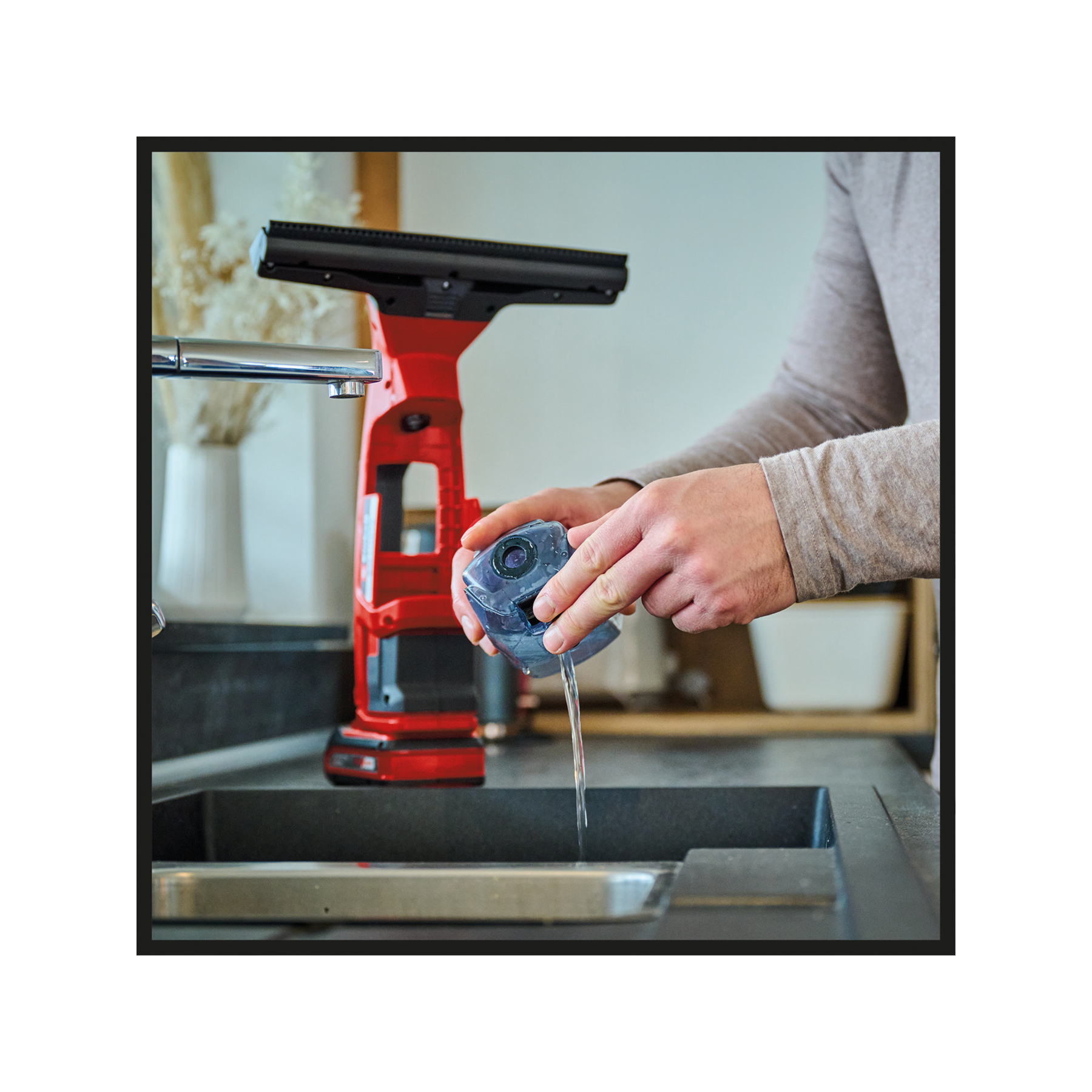 Osoba płucze przezroczysty pojemnik wodą nad zlewem. Po lewej stronie znajduje się czerwono-czarne urządzenie czyszczące.
