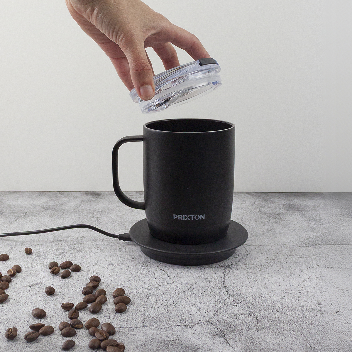 Ręka nakłada pokrywkę na czarny kubek. Kubek stoi na czarnym podgrzewaczu z kablem zasilającym. Ziarna kawy są rozrzucone w pobliżu.