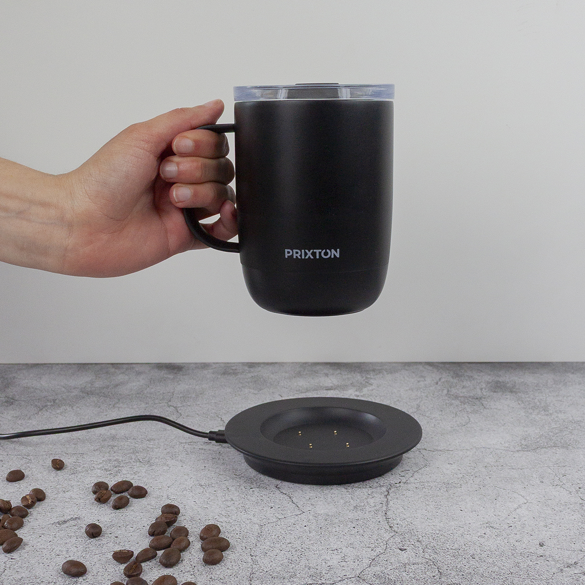 Ręka trzyma czarny kubek PRIXTON nad stacją ładującą. Ziarna kawy rozsiane na szarej powierzchni.