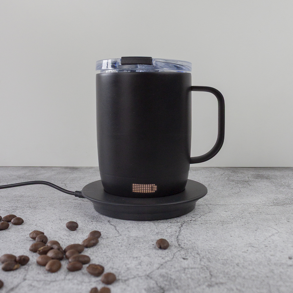 Black mug on a black coaster with coffee beans scattered around. The mug has a handle and a clear lid.  Gray background.