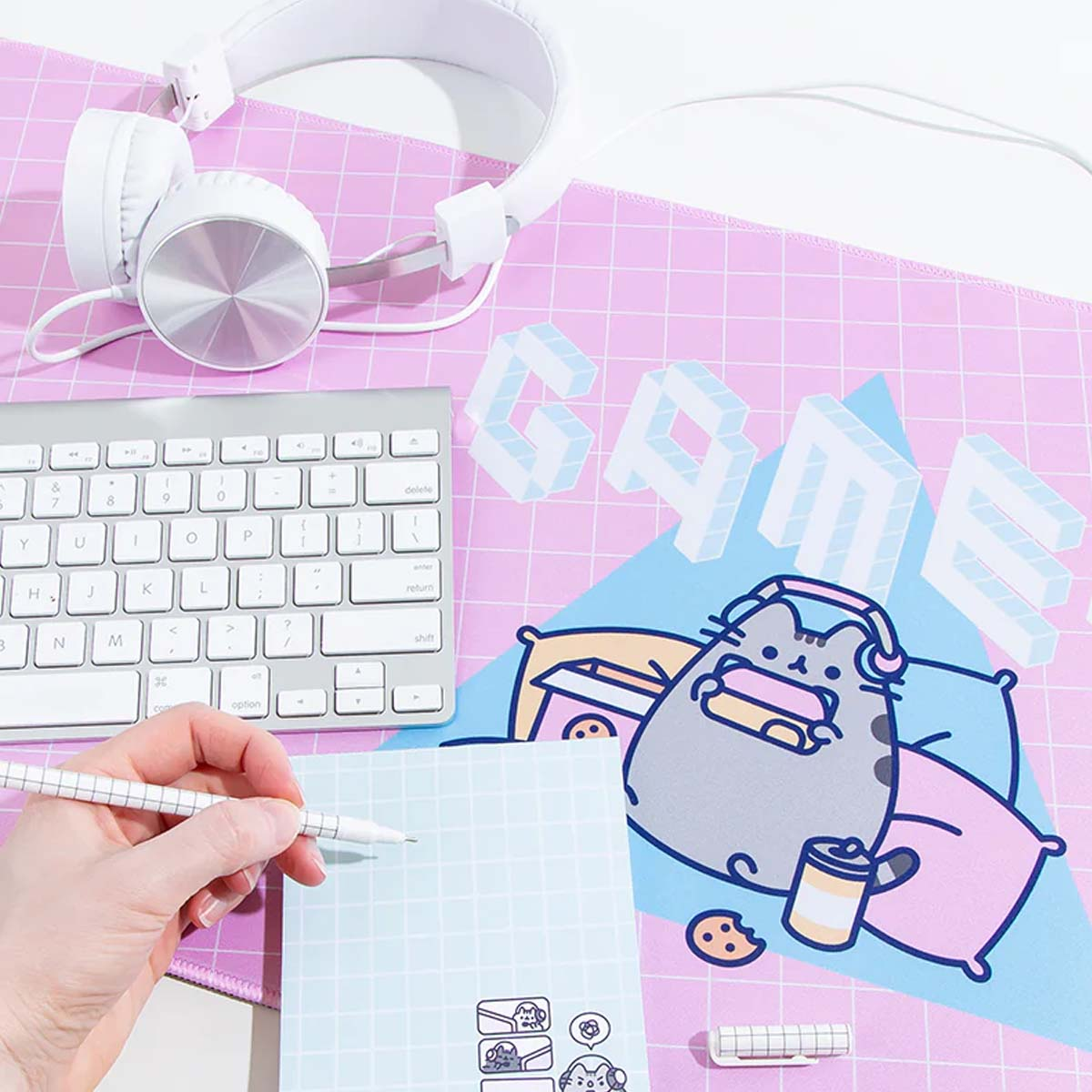 Une configuration de bureau avec un casque, un clavier, un bloc-notes et un tapis de souris Pusheen. Une main tient un stylo au-dessus du bloc-notes.