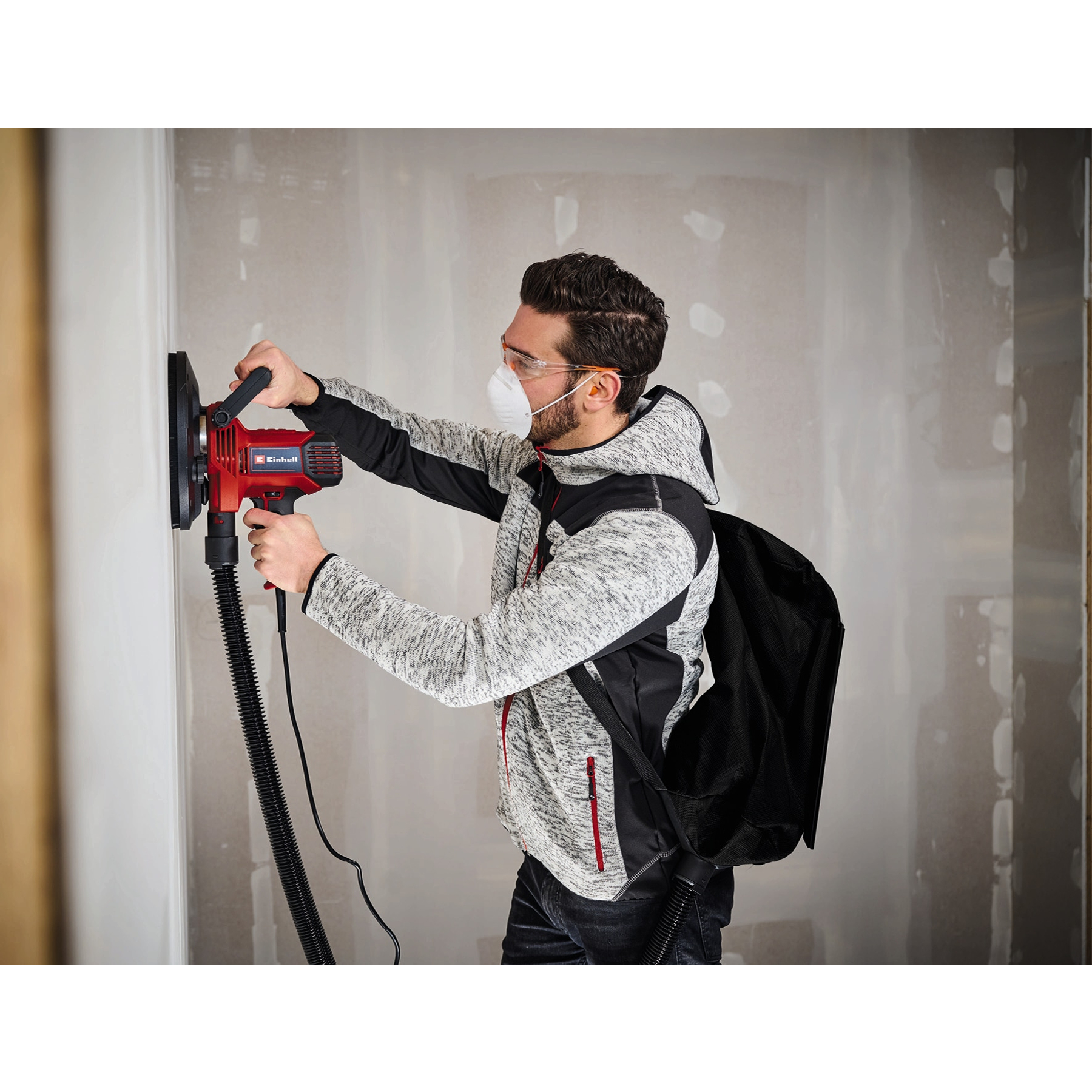 Mann in Schutzkleidung schleift mit einem roten Elektroschleifer eine Wand. Schwarzer Schlauch und Tasche befestigt. Grauer Wandhintergrund.