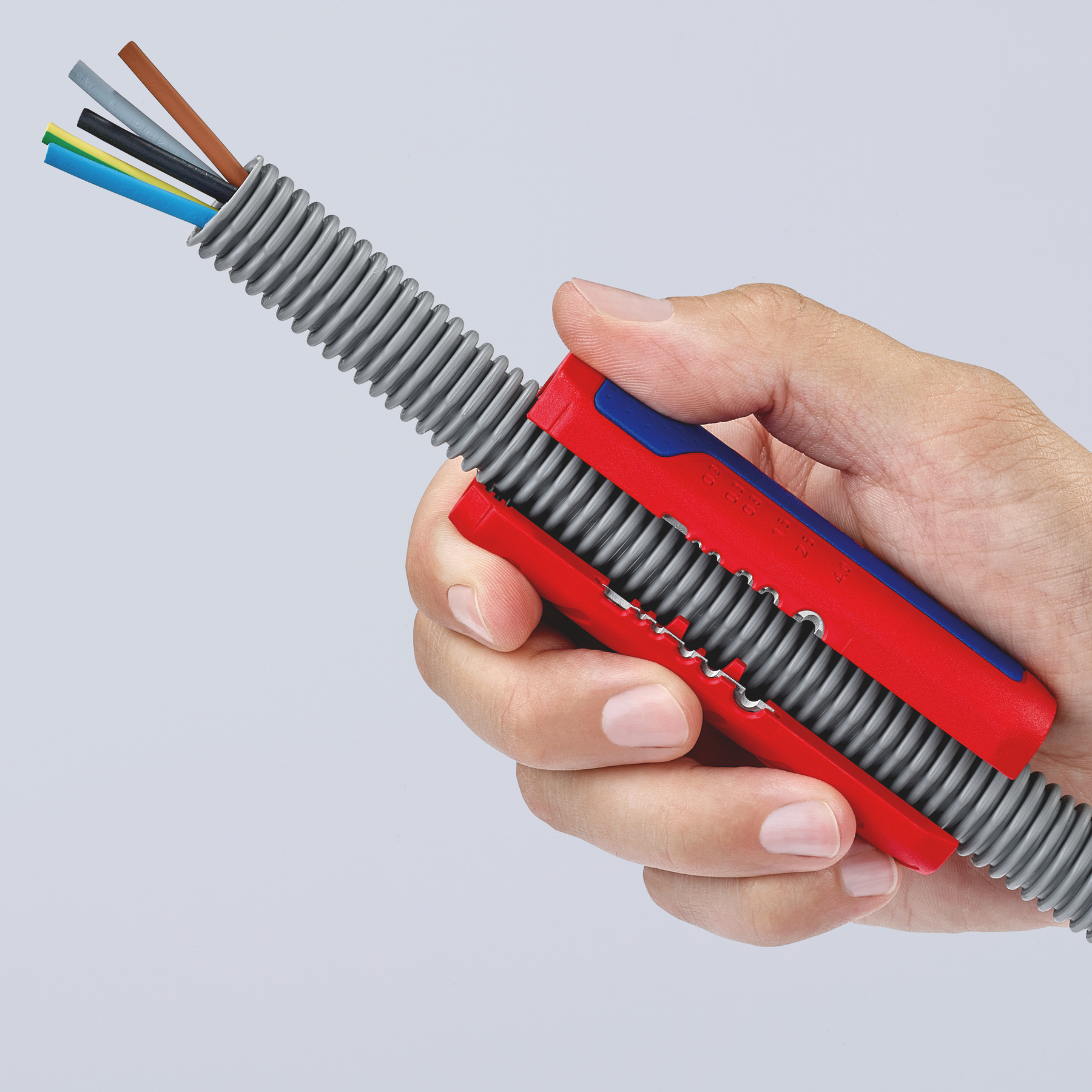 Hand holding a red and blue wire stripper, cutting into a gray corrugated conduit with colored wires protruding.