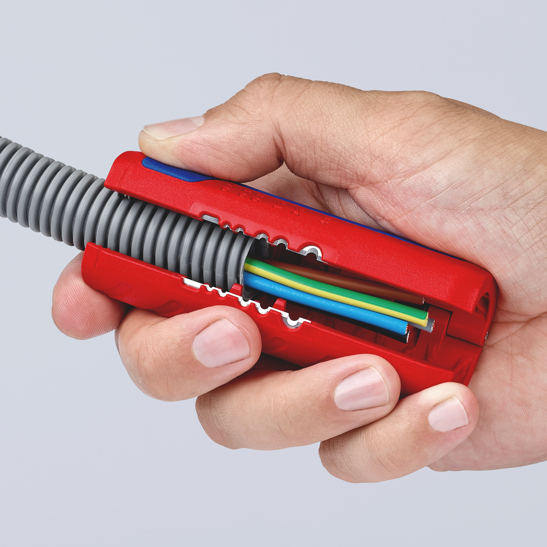 Hand holding a red and blue wire stripper, stripping wires from a gray corrugated tube. Wires are blue, yellow, green, and brown.