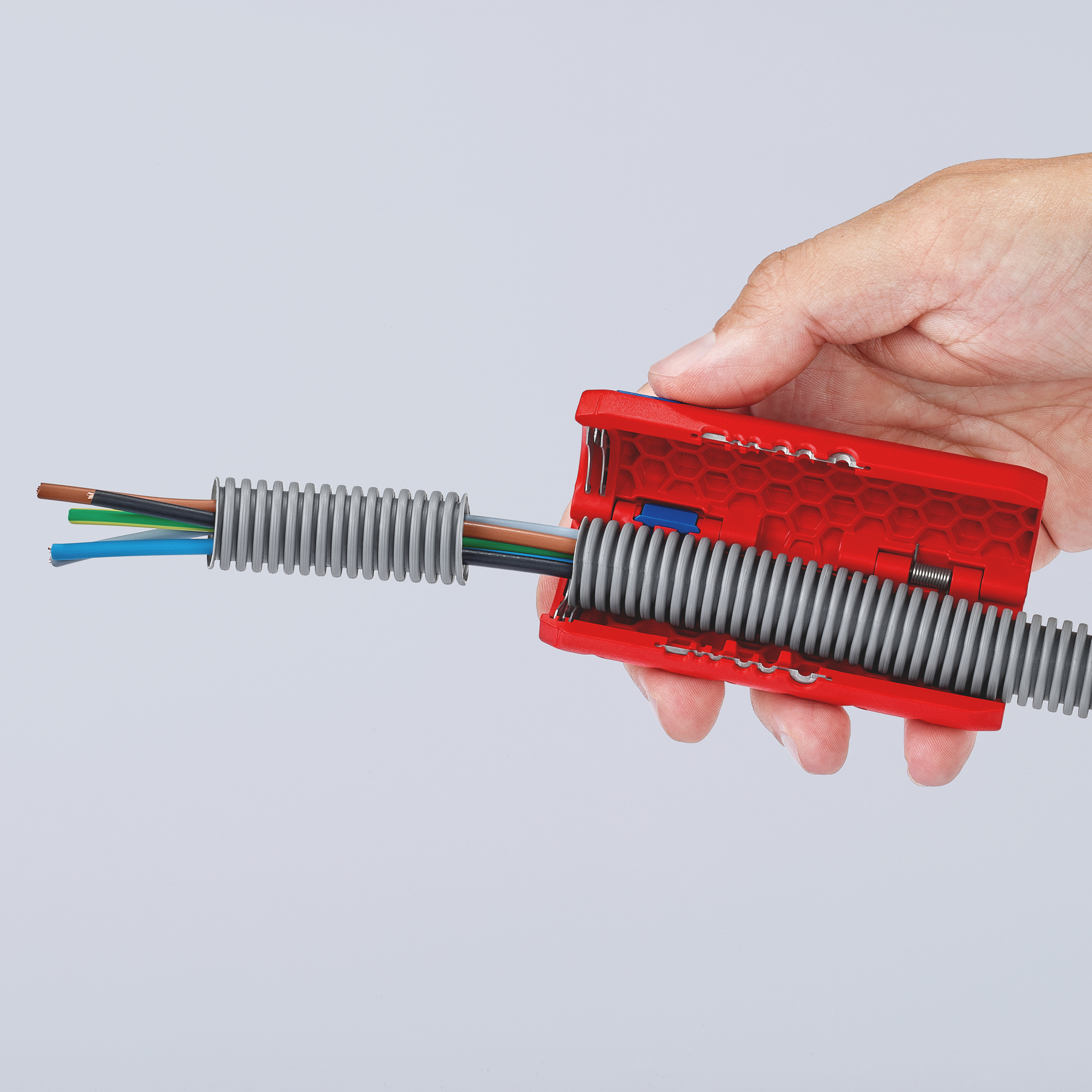 Hand holding a red tool stripping a gray corrugated tube filled with colored wires against a light gray background.