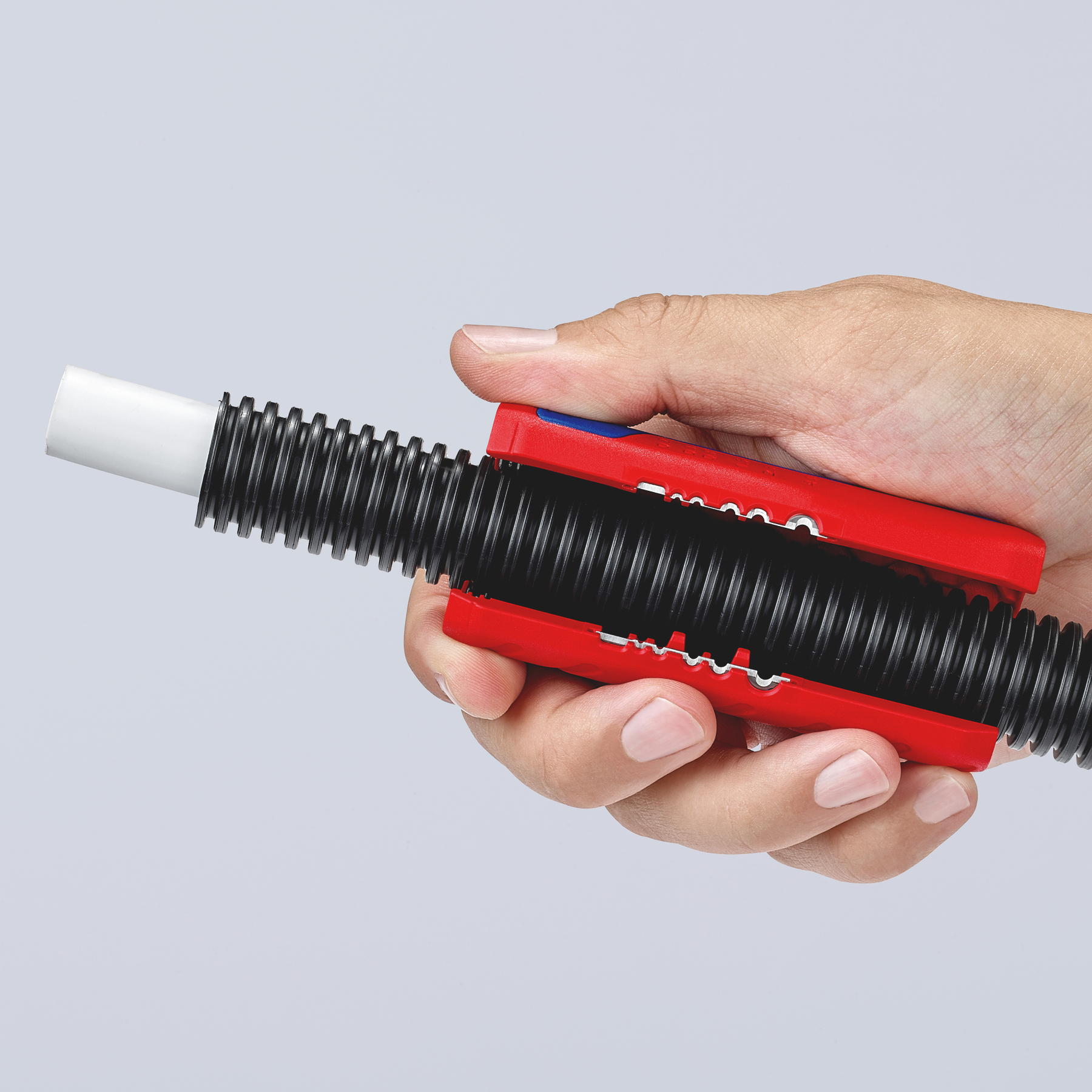 Hand holding a red and blue tool cutting a black corrugated tube, with a white tube inserted. Plain background.