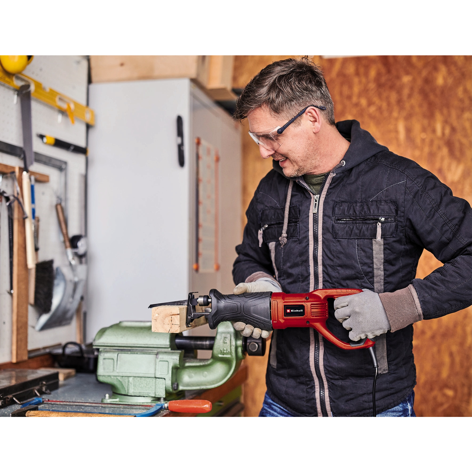 Mężczyzna używa czerwono-czarnej piły do drewna w warsztacie. Ma okulary ochronne i rękawice.