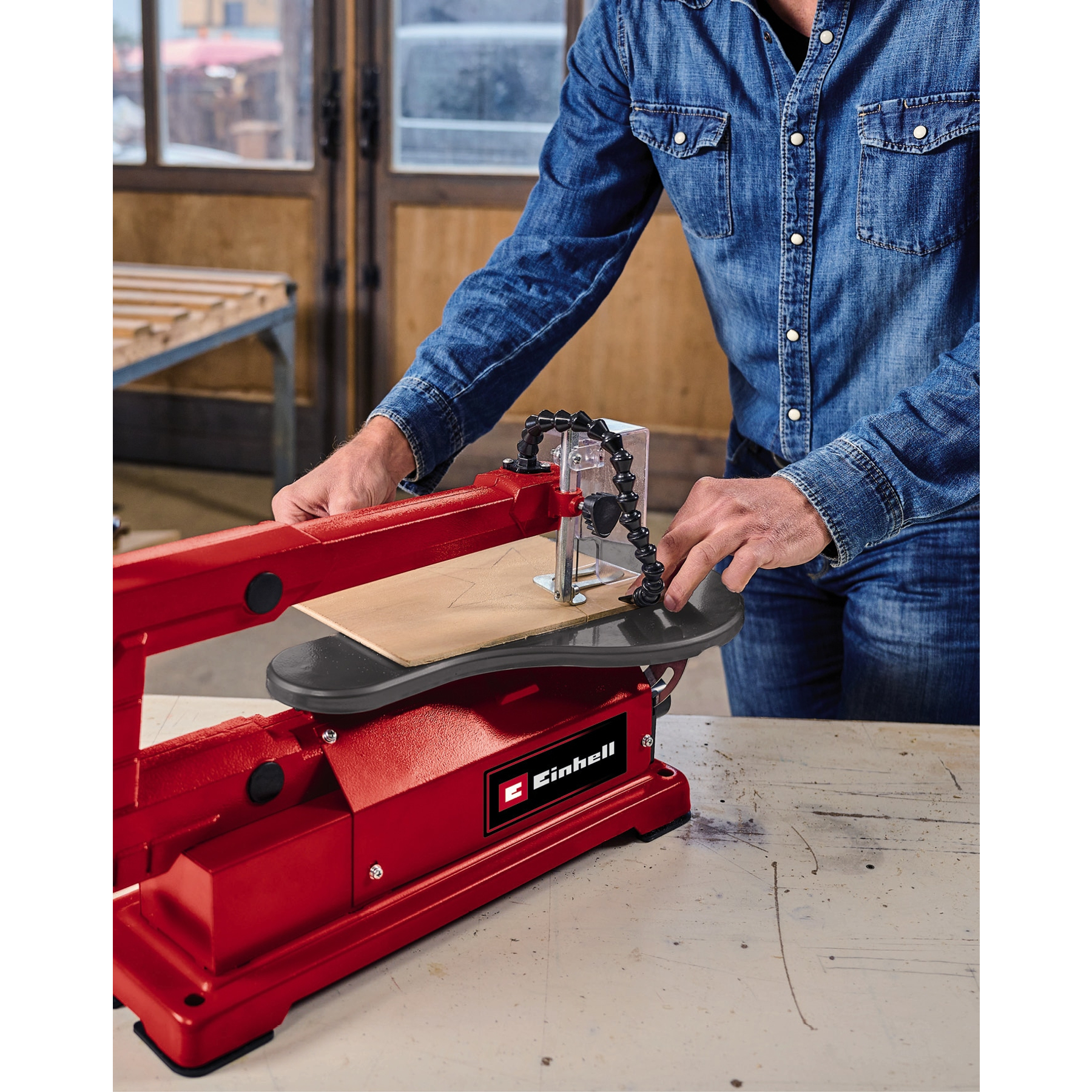 Osoba używa czerwonej wyrzynarki Einhell do cięcia drewnianej deski. Scena rozgrywa się w warsztacie.