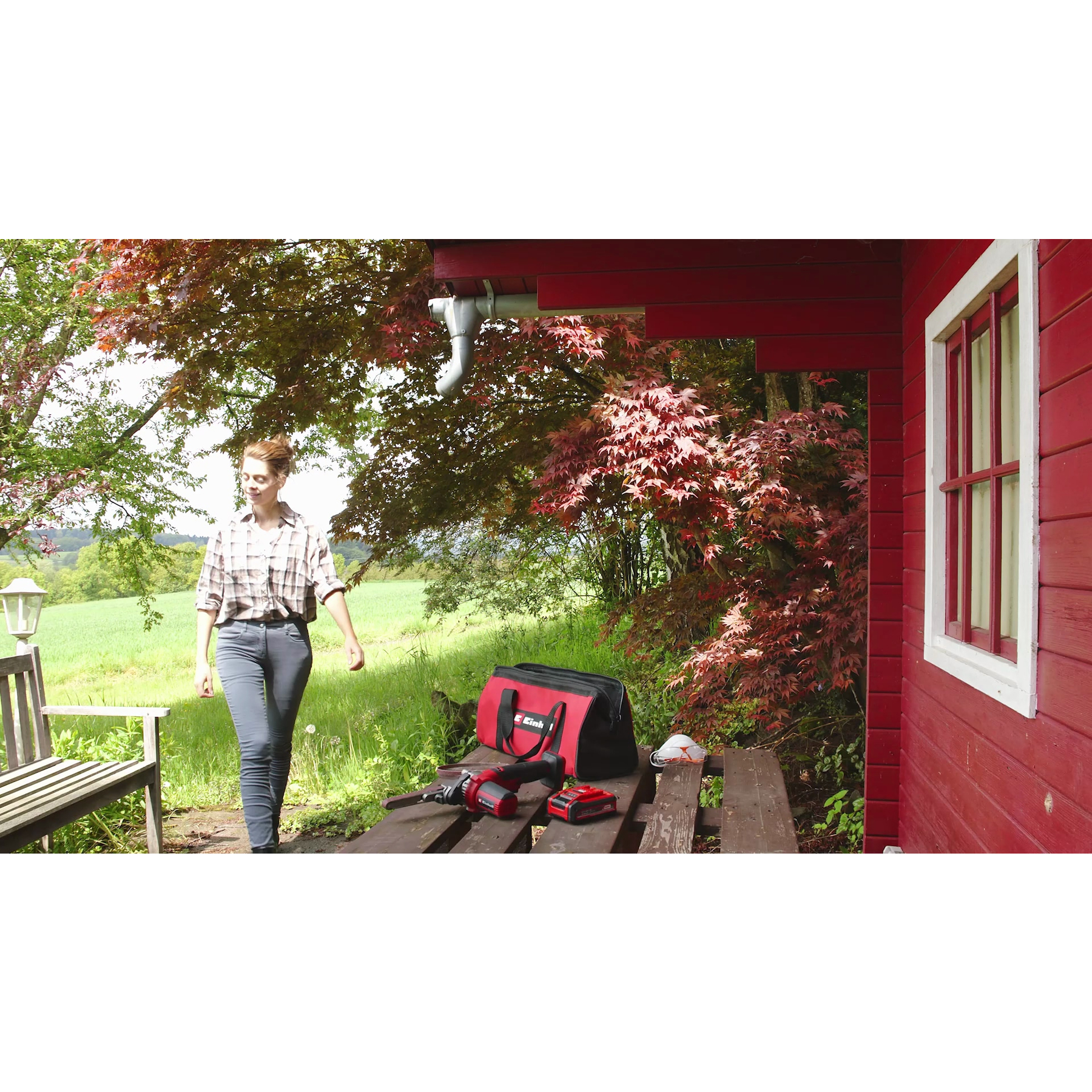 Kobieta przechodzi obok narzędzi na stole w pobliżu czerwonej szopy. Narzędzia obejmują szlifierkę, baterię i torbę. Zielone pole w tle.