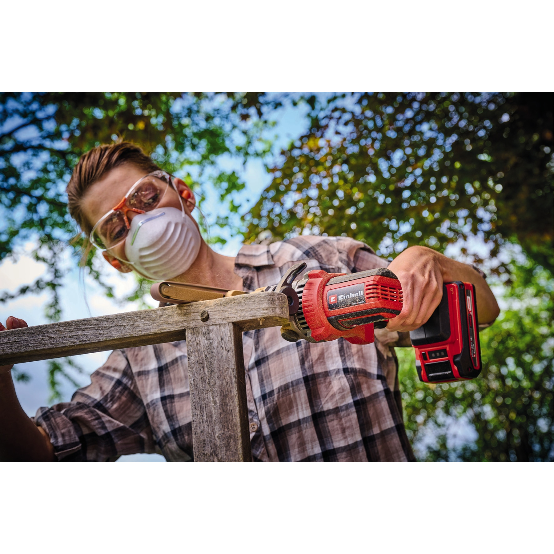 Osoba w masce i okularach używa czerwonego elektronarzędzia na drewnie na zewnątrz. W tle widoczne są drzewa.