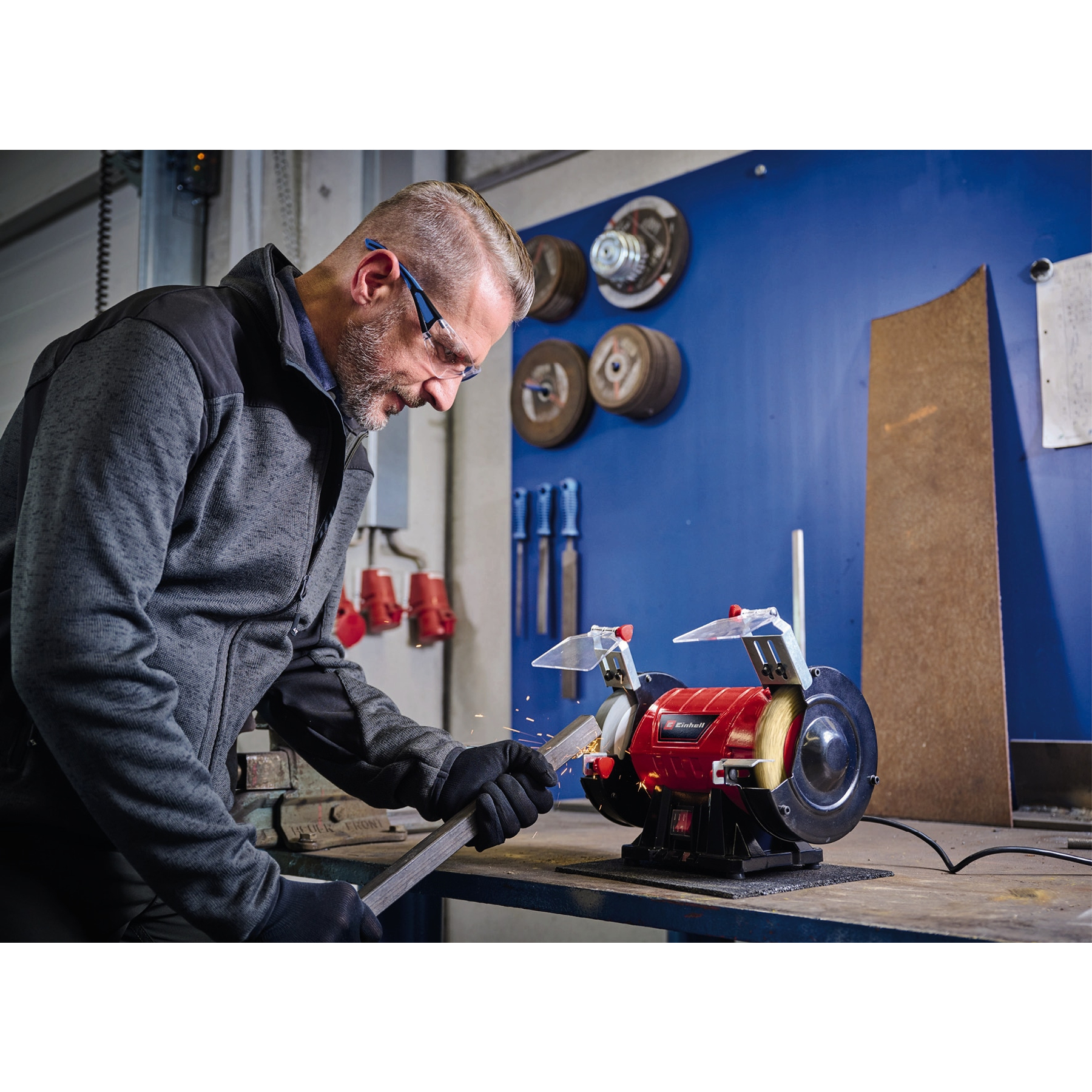 Mężczyzna szlifuje metal maszyną. Iskry lecą. Tło ma narzędzia i niebieską ścianę.