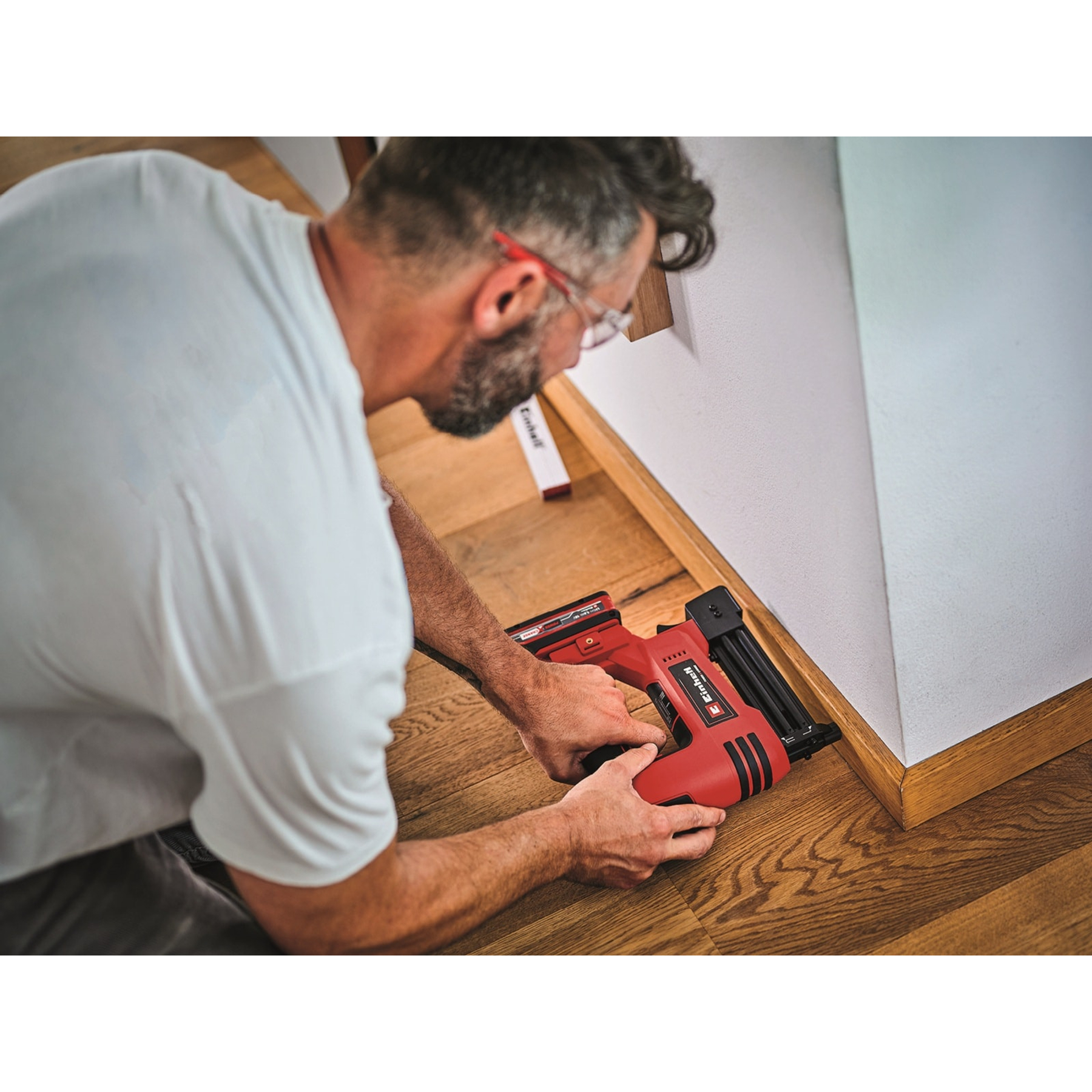 Een man in een wit shirt gebruikt een rood spijkerpistool op een houten vloer naast een muur.