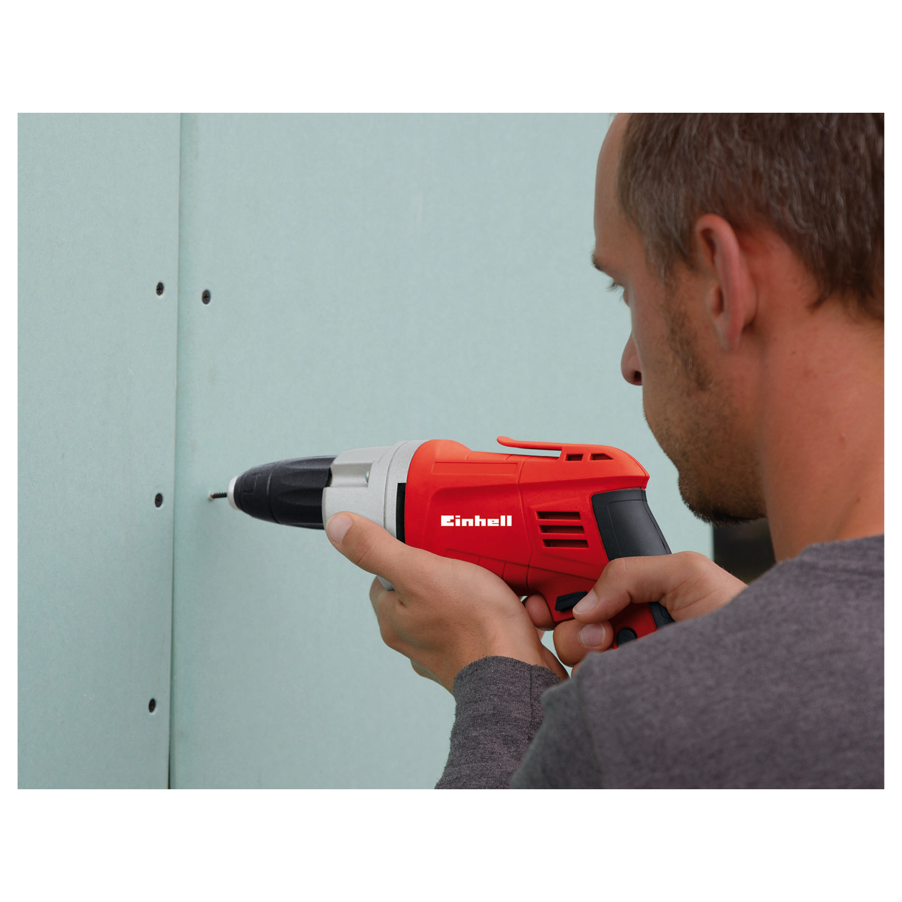 Ein Mann bohrt mit einem rot-grauen Einhell-Bohrer in eine Wand. Die Wand ist hellgrün.