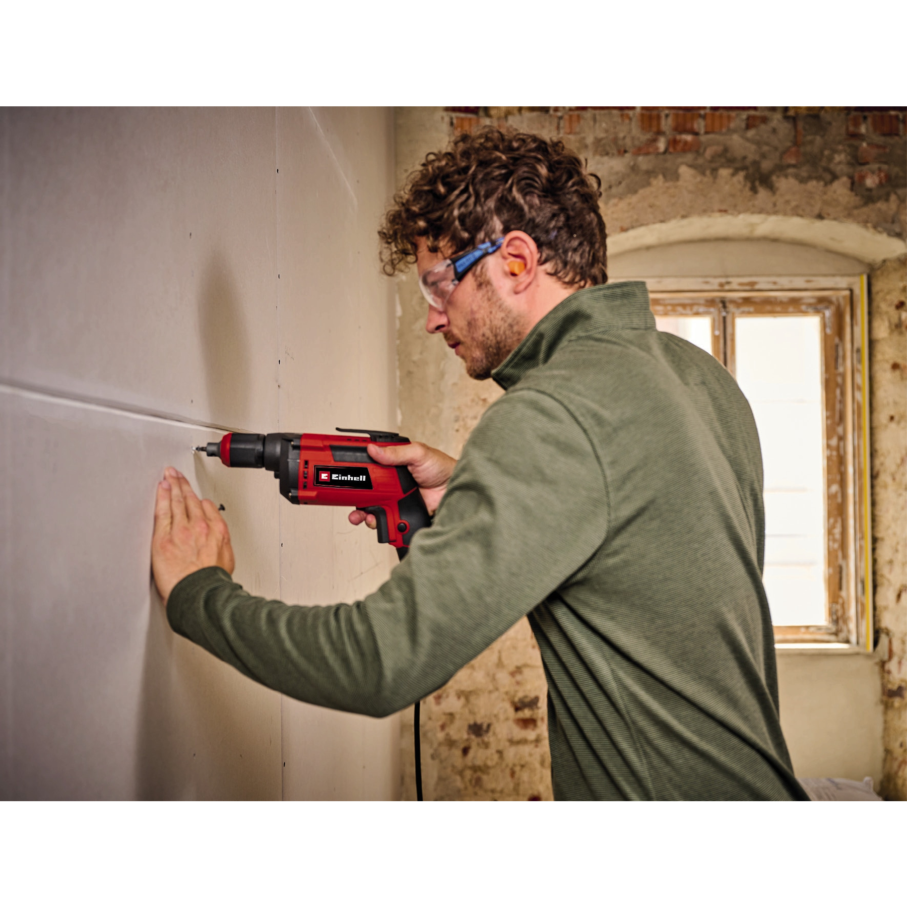 Ein Mann bohrt in eine Wand. Er trägt Schutzbrille, Ohrstöpsel und ein grünes Hemd. Der Bohrer ist rot und schwarz.