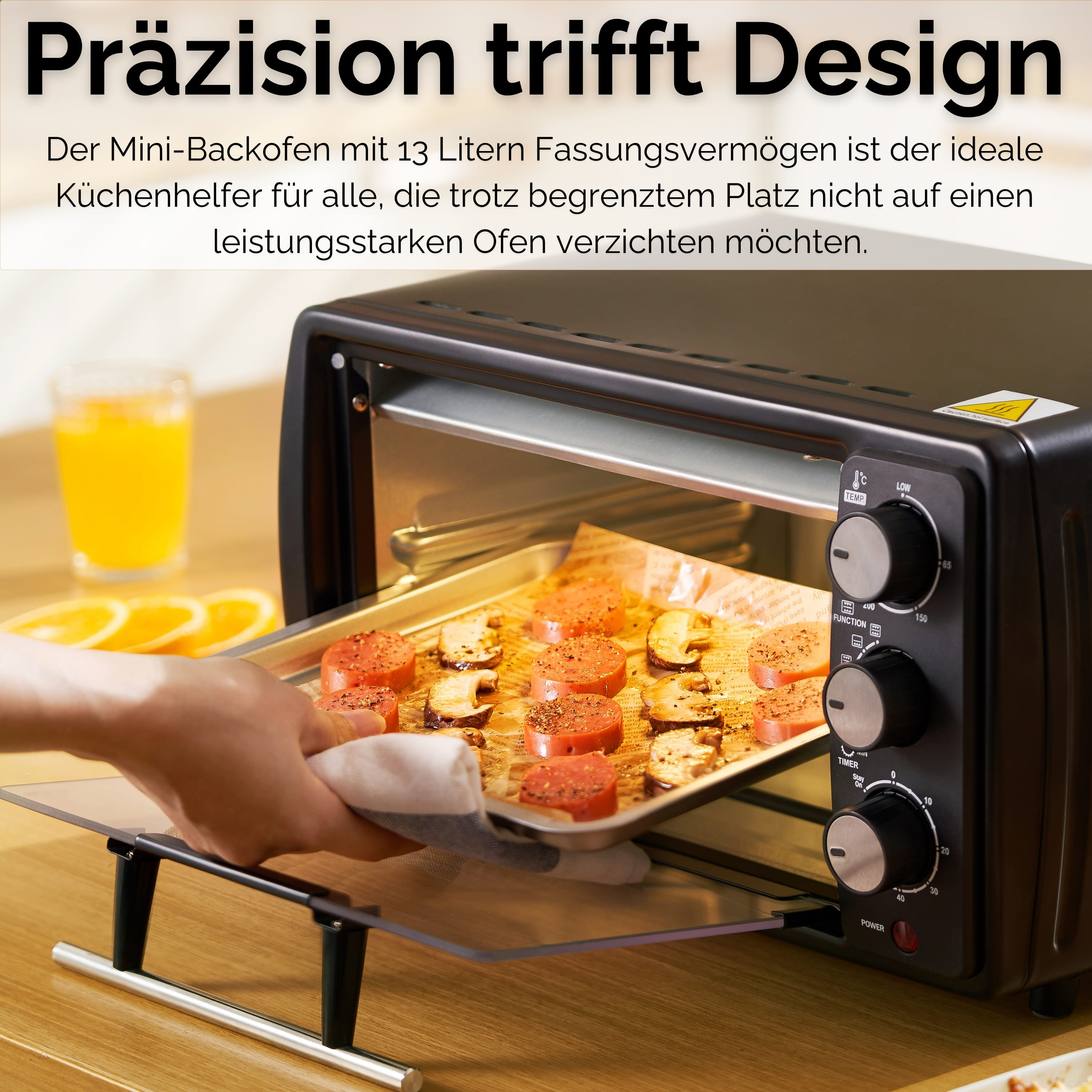 Ein schwarzer Mini-Backofen mit Essen auf einem Tablett, das herausgenommen wird. Ein Glas Orangensaft steht im Hintergrund.