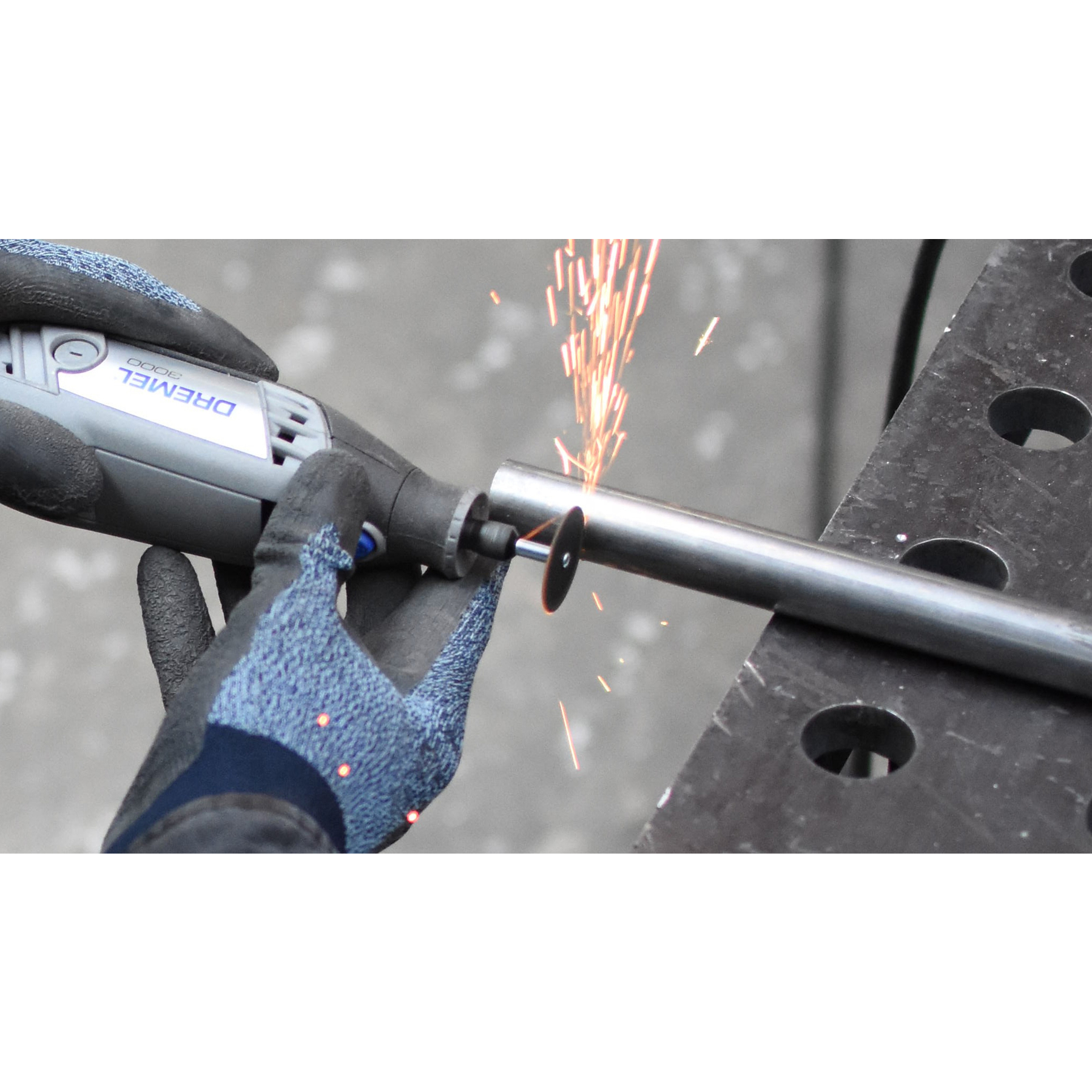 Eine Person schneidet ein Metallrohr mit einem Dremel-Werkzeug. Funken fliegen. Graue Handschuhe und eine schwarze Metalloberfläche sind sichtbar.