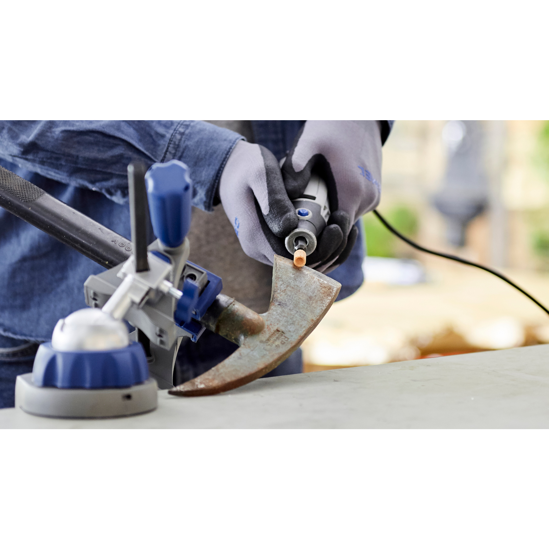 Eine Person schleift eine rostige Axt mit einem Rotationswerkzeug. Graue Handschuhe, blaue und graue Werkzeuge und ein Jeanshemd sind sichtbar.